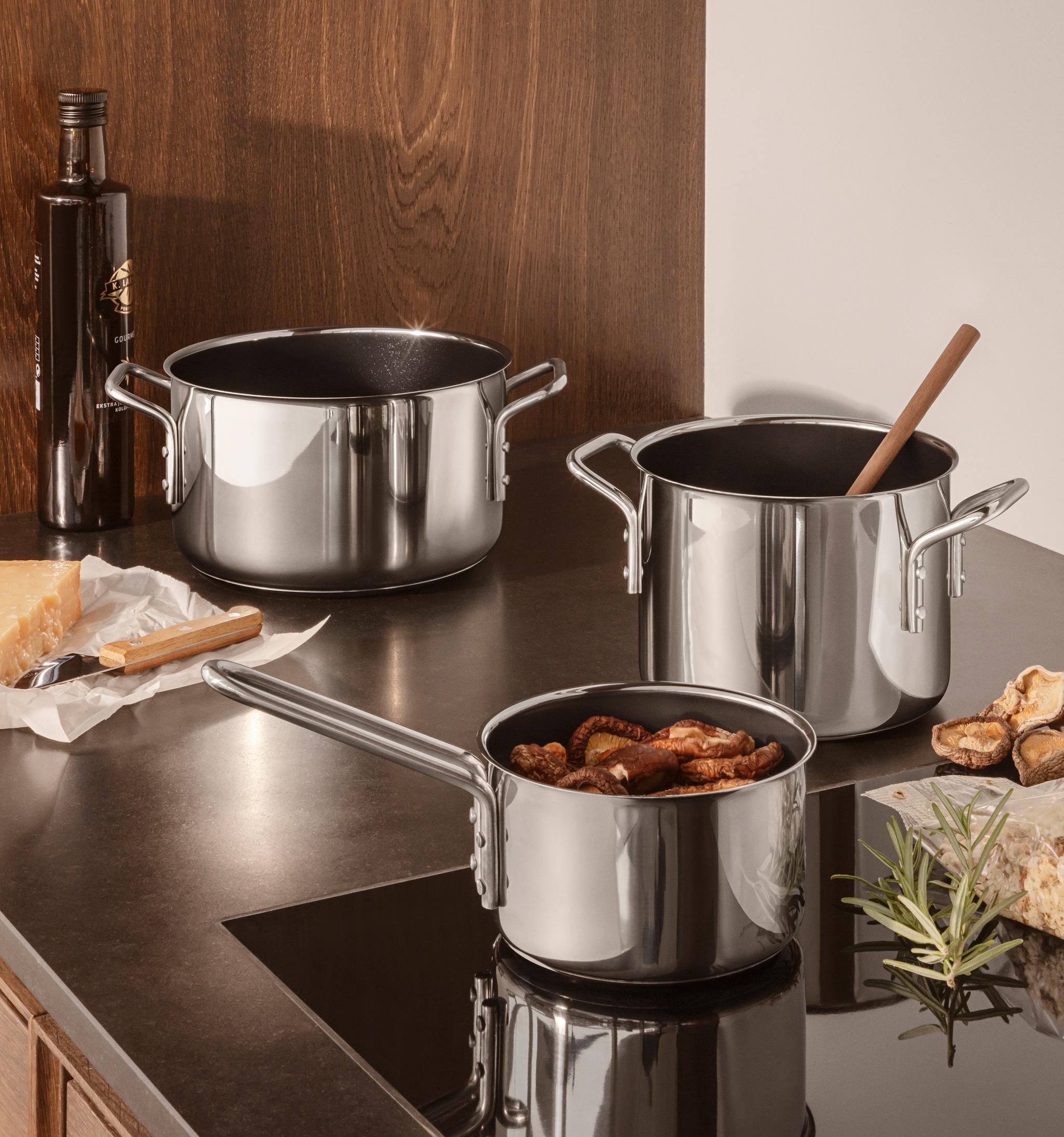 Kaksi ruostumattomasta teräksestä valmistettua kattilaa ja kattila, jossa on sieniä tummalla keittiön tasolla, juustoa, oliiviöljyä ja rosmariinia.