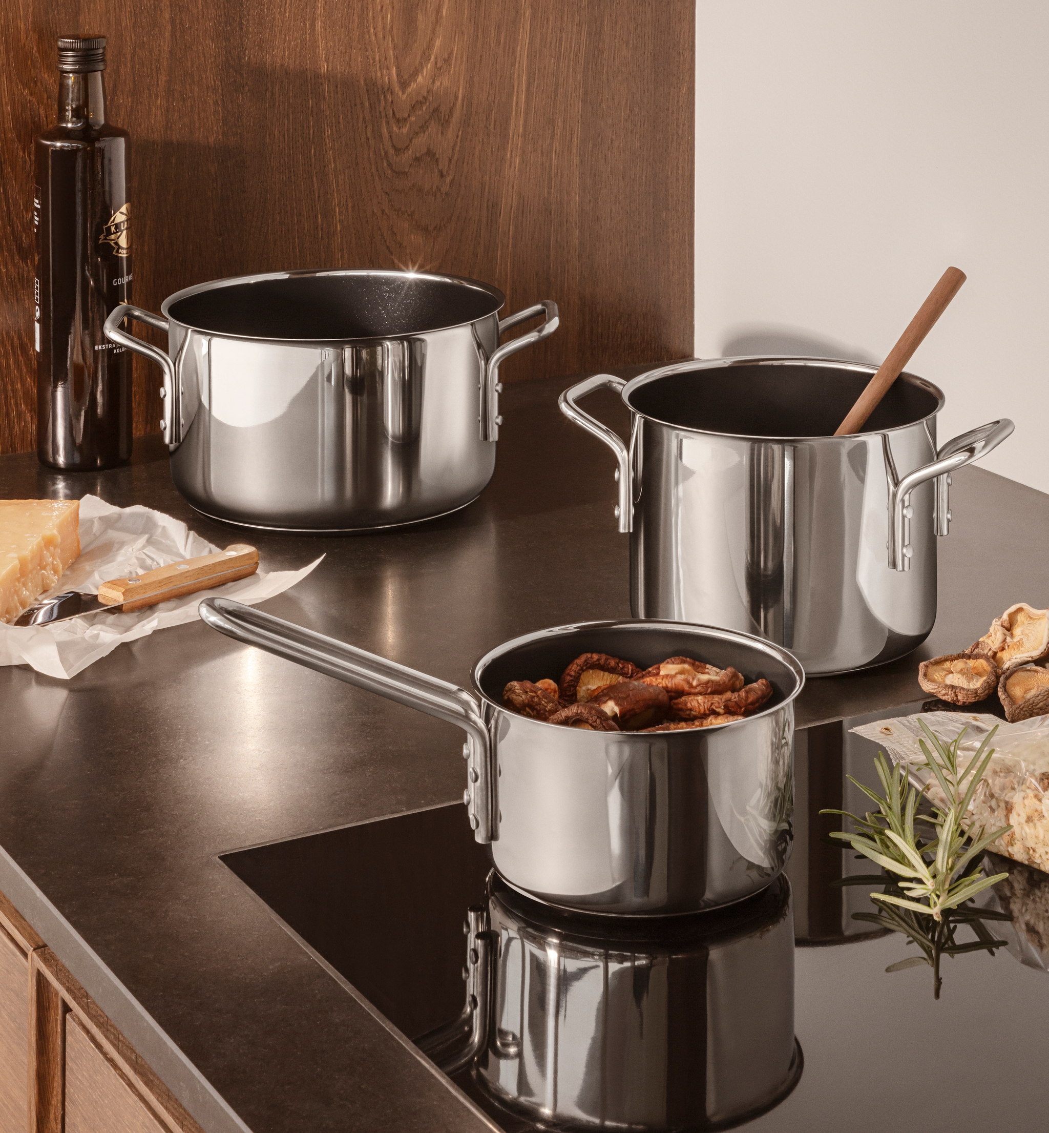 Kolme ruostumattomasta teräksestä valmistettua kattilaa keittiön tiskillä, jossa on juustoa, oliiviöljyä ja kuivattuja sieniä.