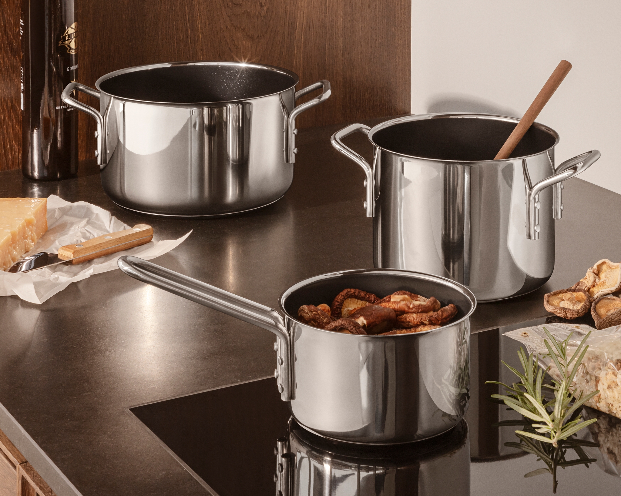 Kaksi ruostumattomasta teräksestä valmistettua kattilaa ja kattila, jossa on sieniä tummalla keittiön tasolla, juustoa, oliiviöljyä ja rosmariinia.