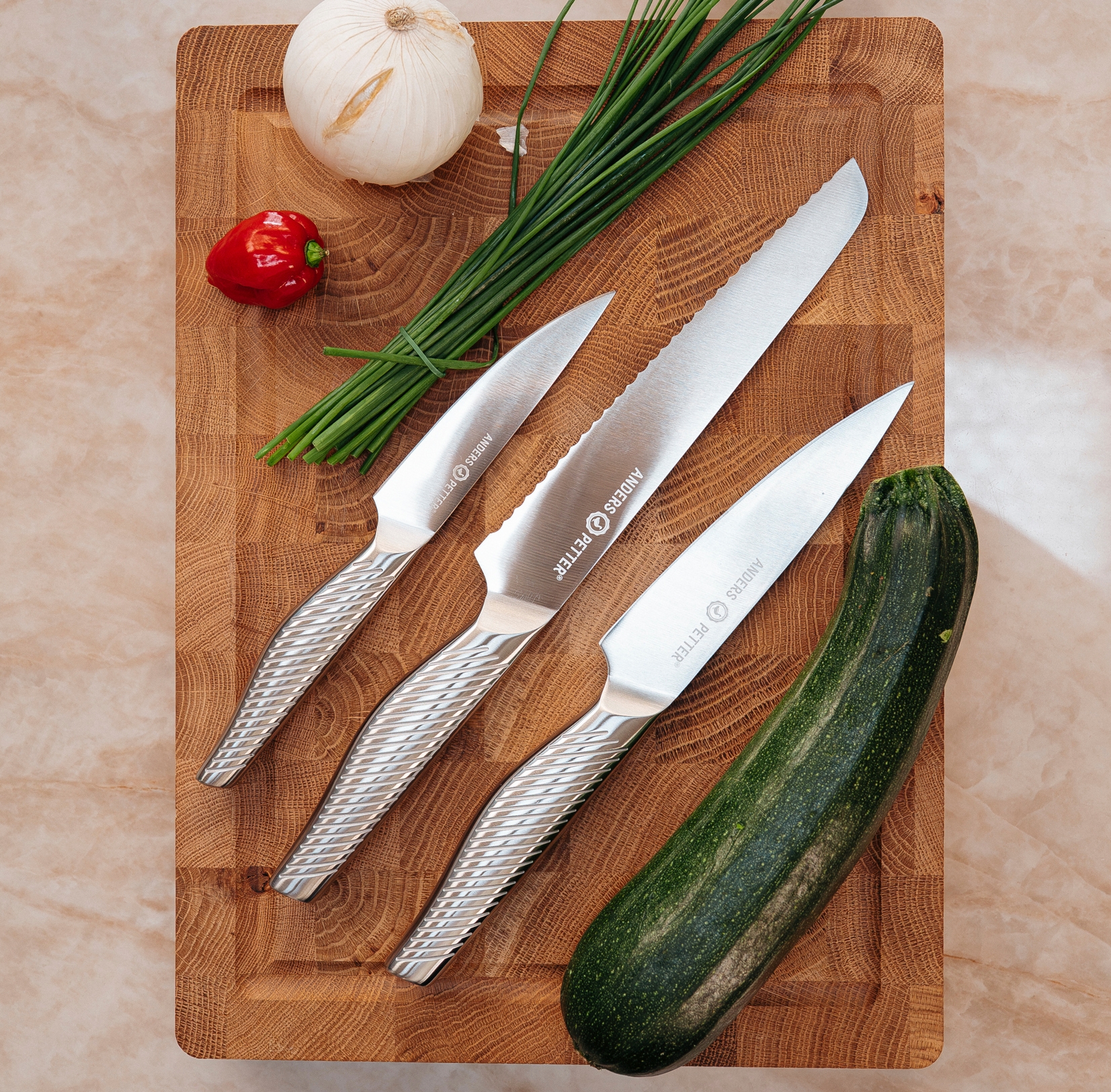 Tre eleganta köksknivar ligger på en träskärbräda tillsammans med zucchini, lök, gräslök och en röd chilipeppar.