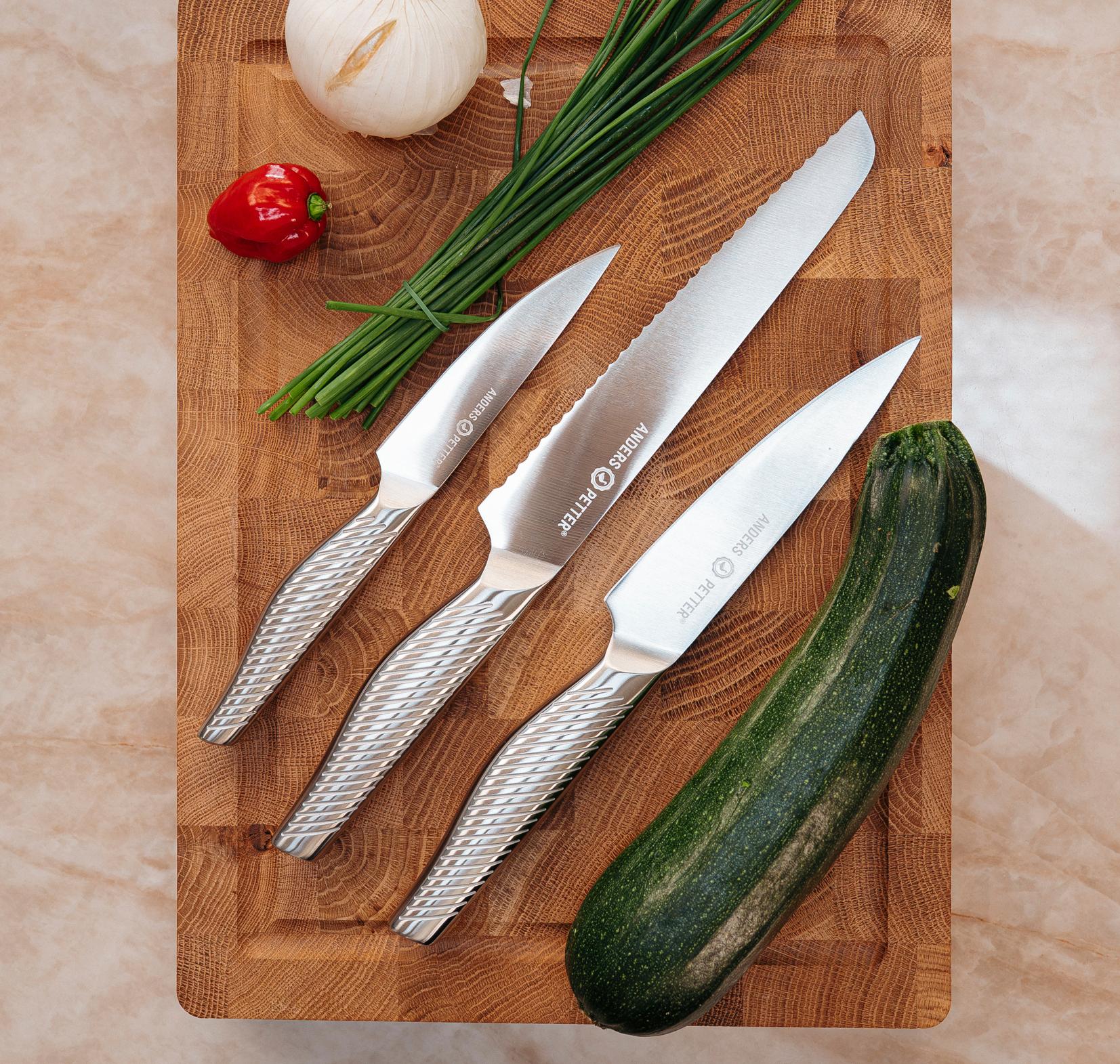 Tre professionella köksknivar ligger på en träskärbräda omgivna av zucchini, lök, gräslök och en röd chilipeppar.