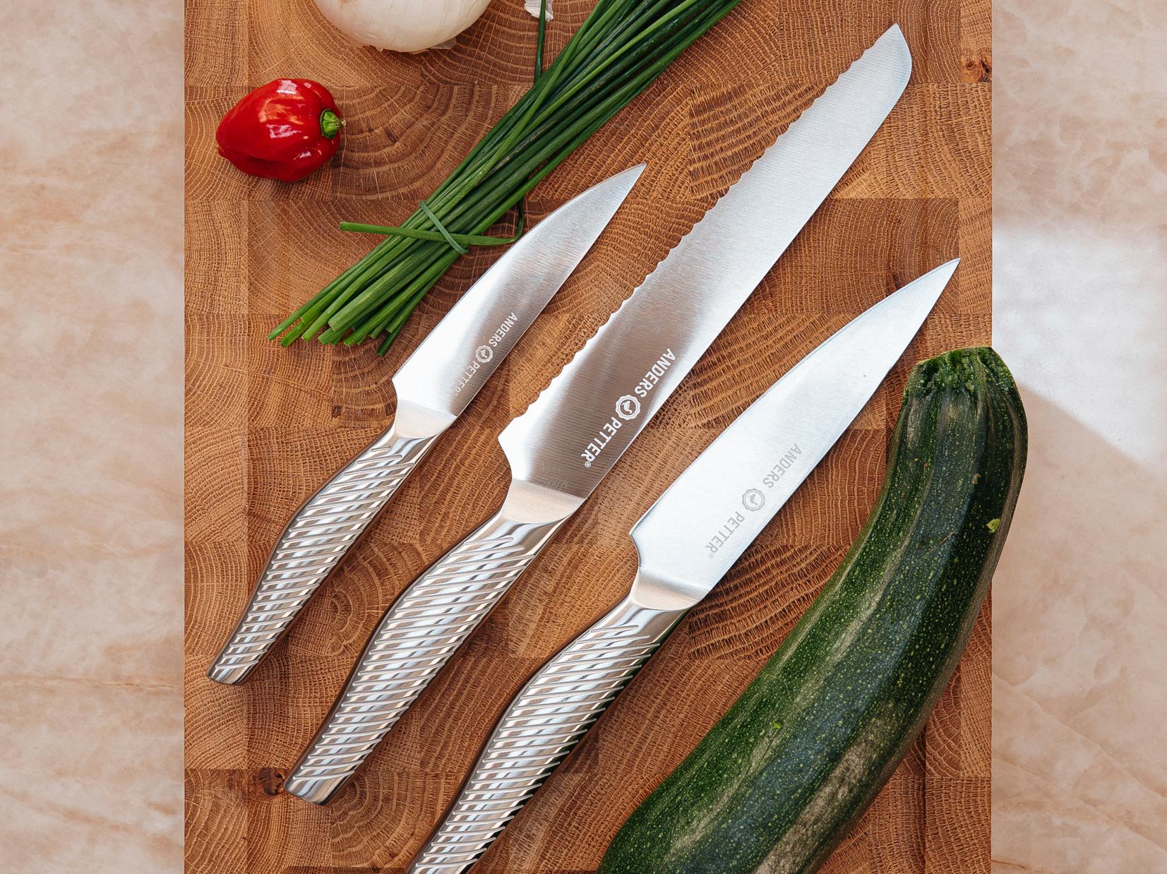 Tre eleganta köksknivar ligger på en träskärbräda tillsammans med zucchini, lök, gräslök och en röd chilipeppar.