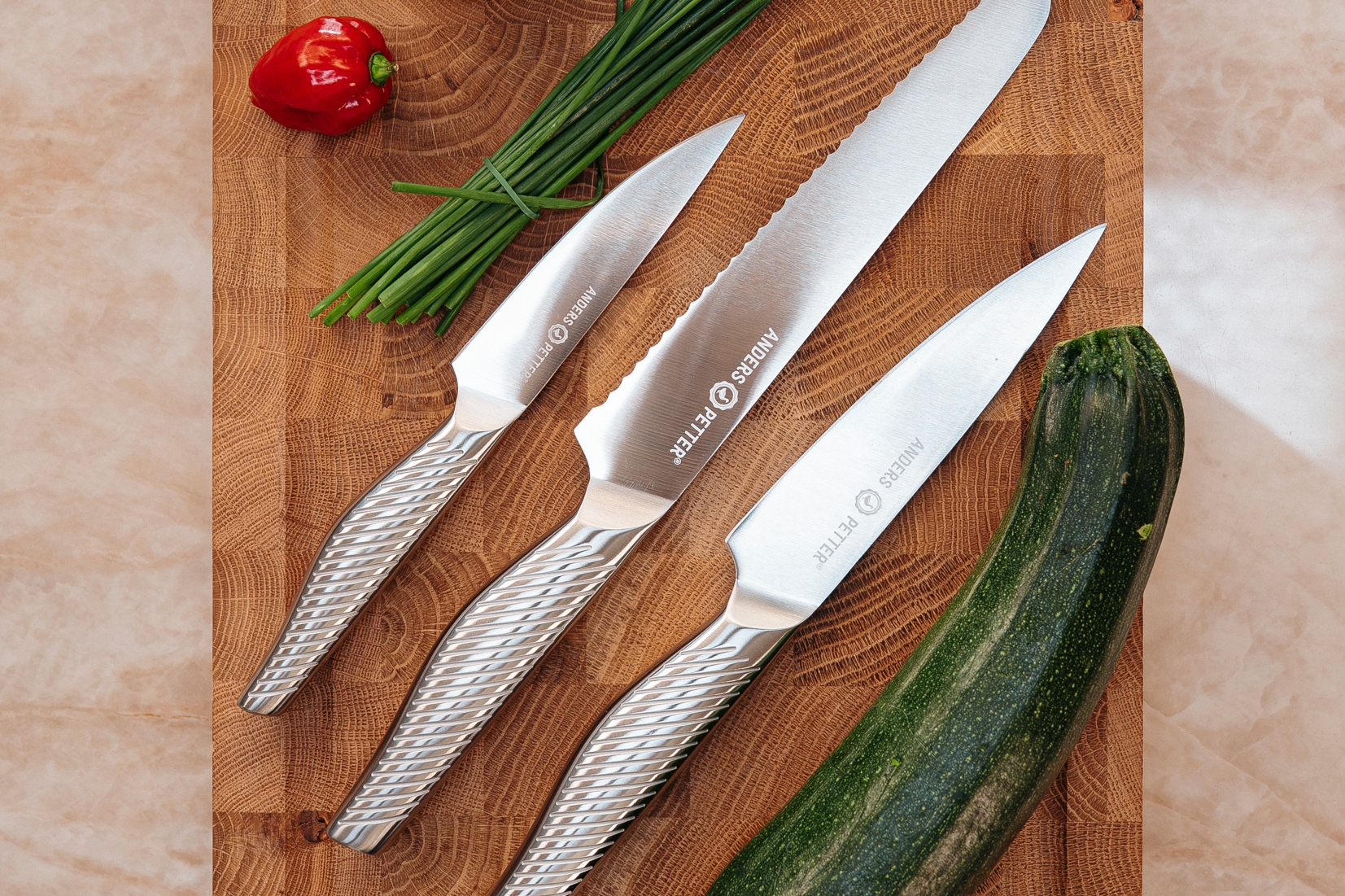 Tre eleganta köksknivar ligger på en träskärbräda tillsammans med zucchini, lök, gräslök och en röd chilipeppar.
