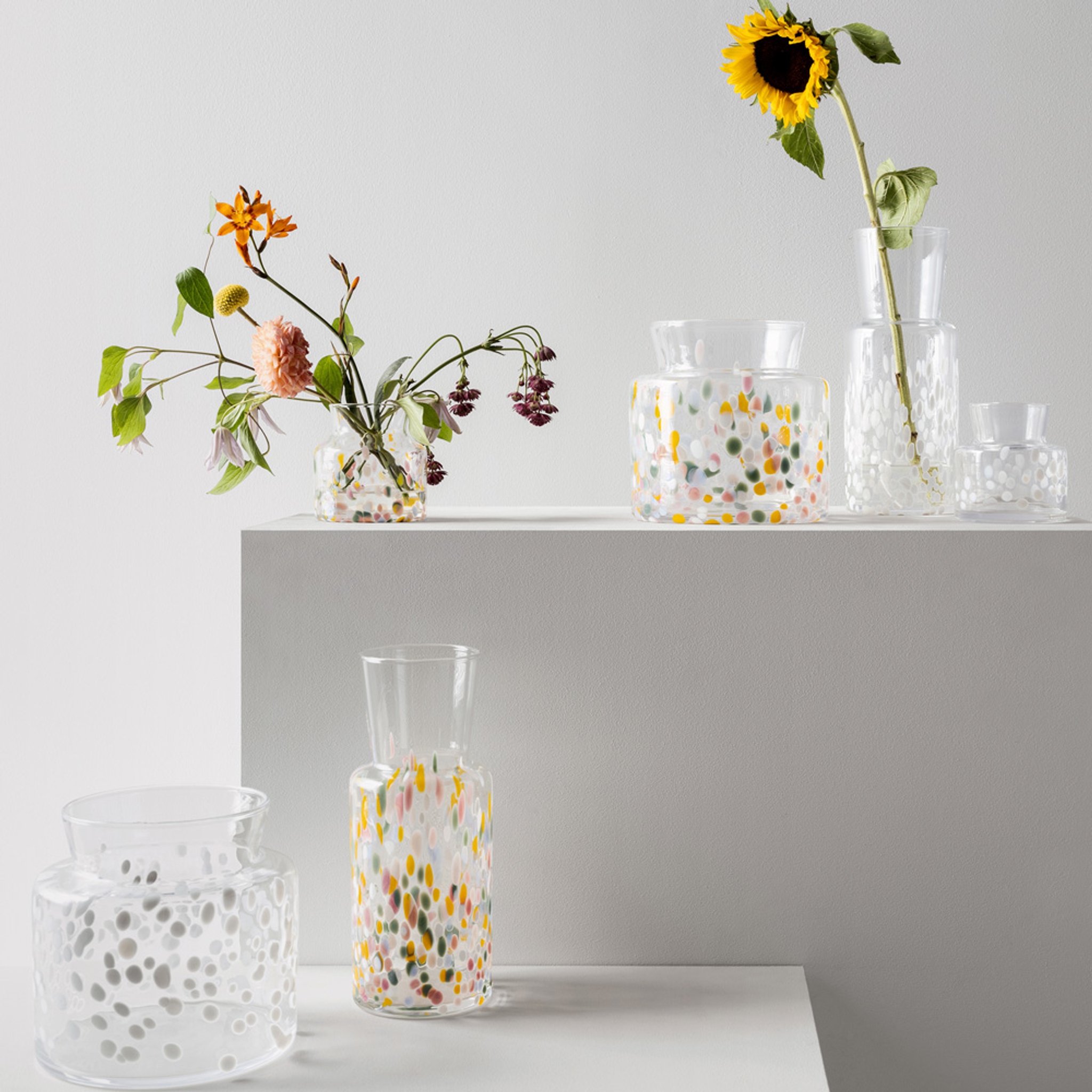 Handgjorda glasvaser med färgglada prickar och sommarblommor mot en ljusgrå bakgrund.