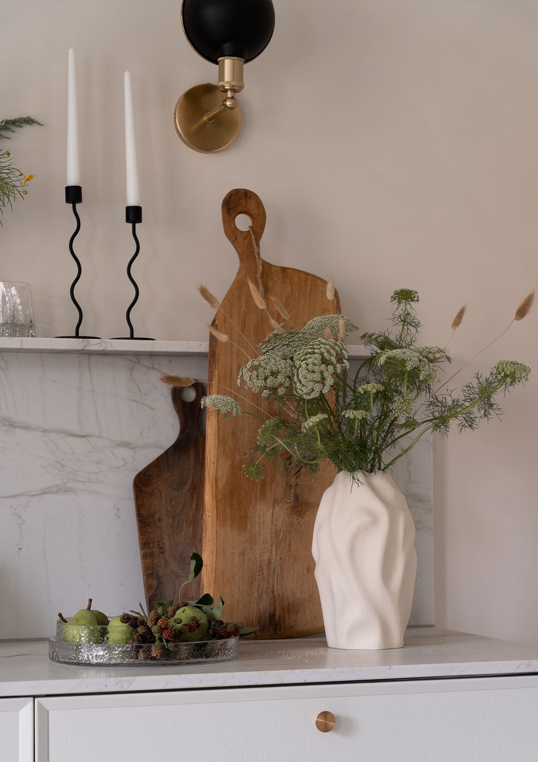 A kitchen counter vignette with a textured white vase of wildflowers, two wooden cutting boards, wavy black candlesticks, and a glass tray of pears and berries.