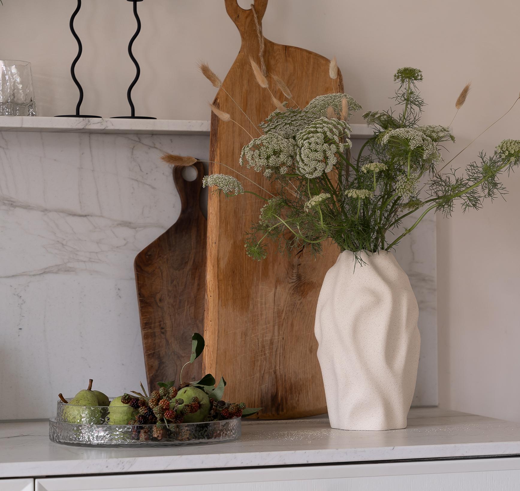 A kitchen counter vignette with a textured white vase of wildflowers, two wooden cutting boards, wavy black candlesticks, and a glass tray of pears and berries.
