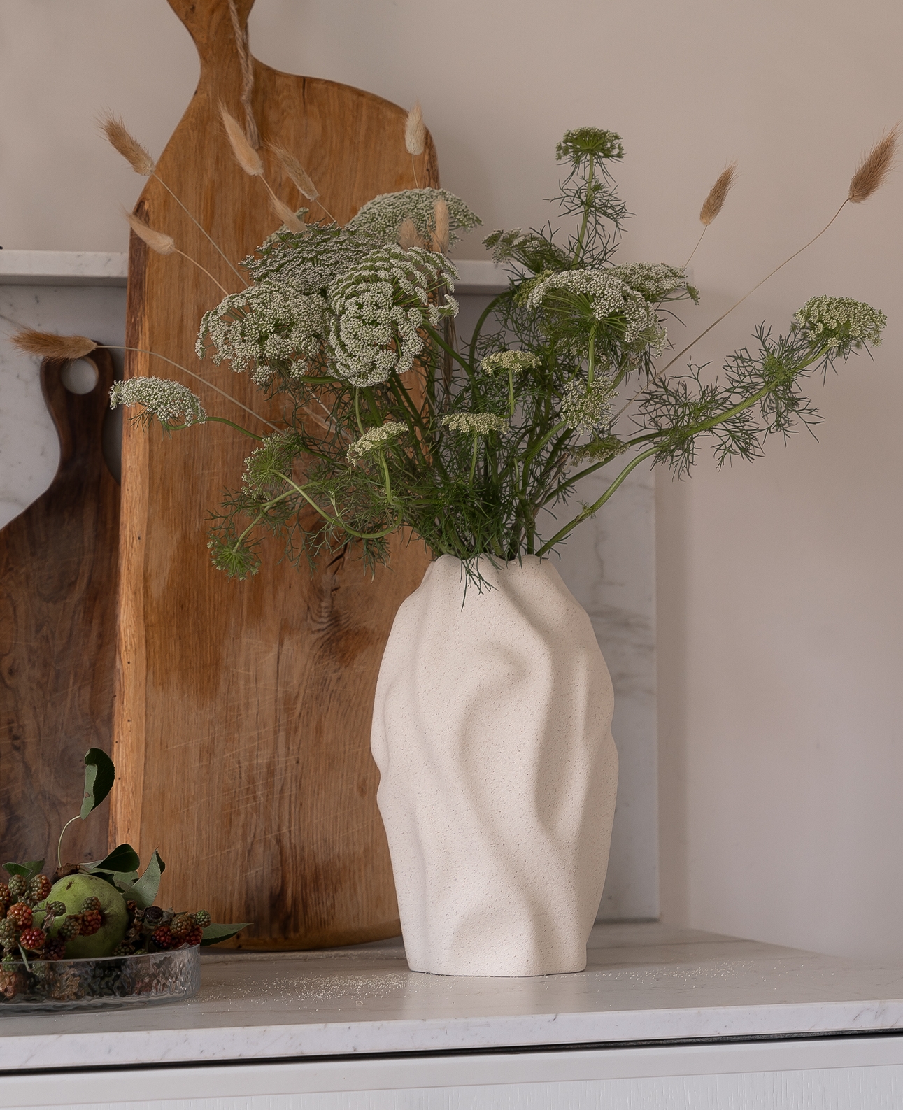 Kitchen counter with black wavy candlesticks, wooden cutting boards, a white vase of Queen Anne's Lace, and a glass bowl of fruit.