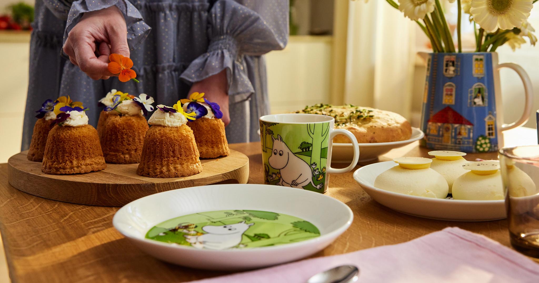 En person dekorerar små kakor med ätbara blommor på ett träbord med Muminporslin, andra desserter och en vas med vita blommor.