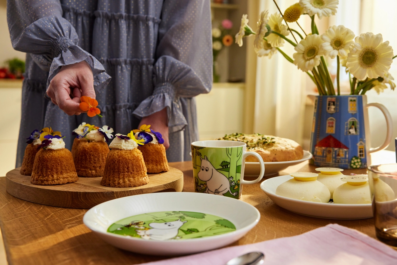 En person dekorerer små kaker med spiselige blomster på et bord fylt med forskjellige desserter og Mummi-tema servise.