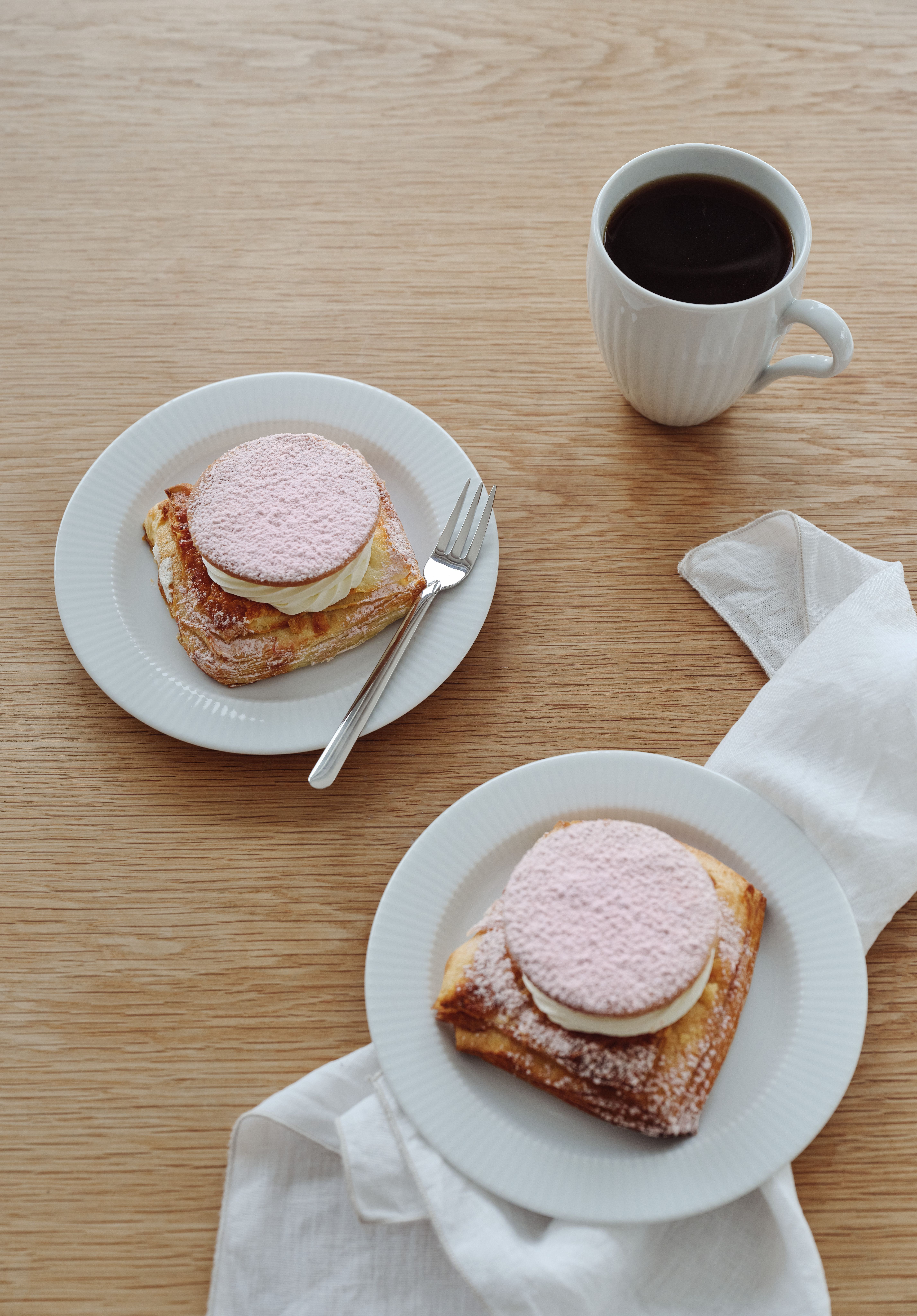 Kaksi neliönmuotoista leivonnaista, joiden päällä on kermaa ja vaaleanpunaisia ​​puuteroituja kiekkoja, valkoisilla lautasilla, kahvikuppi ja lautasliina puupöydällä.