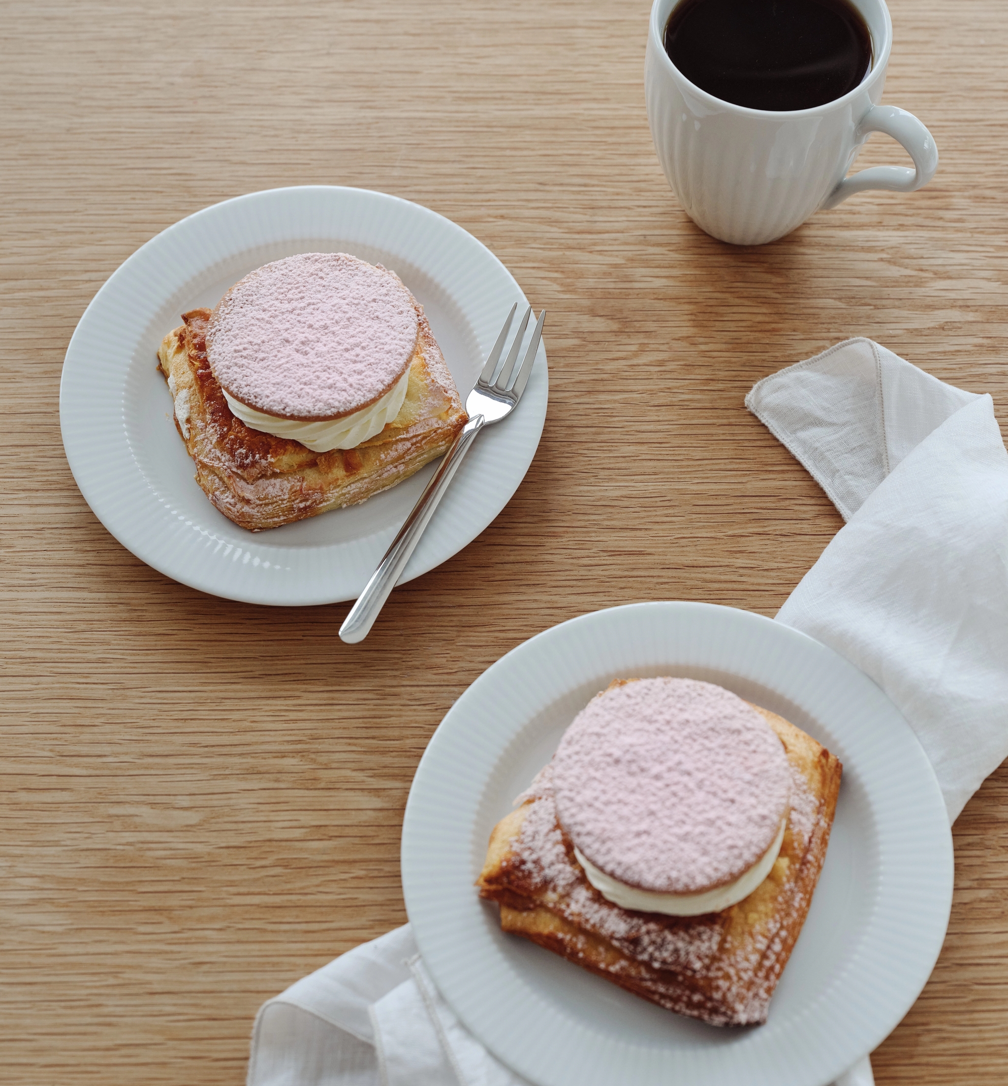 Kaksi neliönmuotoista leivonnaista, joiden päällä on kermaa ja vaaleanpunaisia puuteroituja kiekkoja, valkoisilla lautasilla, kahvikuppi ja lautasliina puupöydällä.