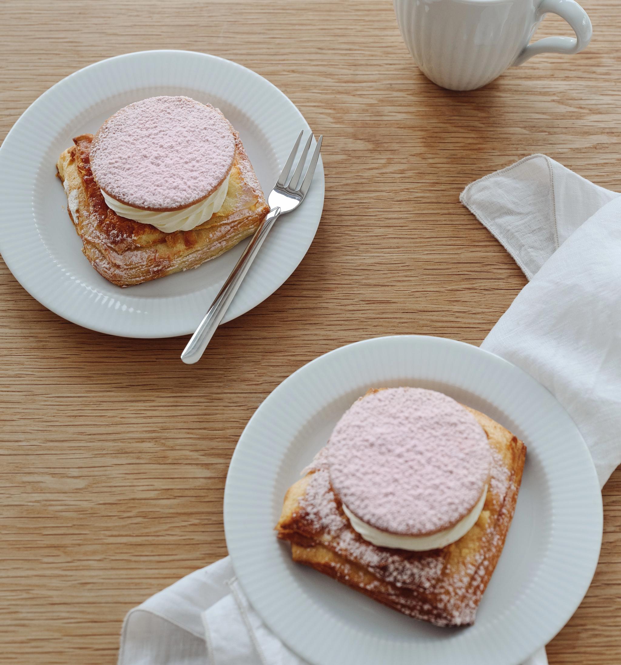 Kaksi neliönmuotoista leivonnaista, joiden päällä on kermaa ja vaaleanpunaisia puuteroituja kiekkoja, valkoisilla lautasilla, kahvikuppi ja lautasliina puupöydällä.