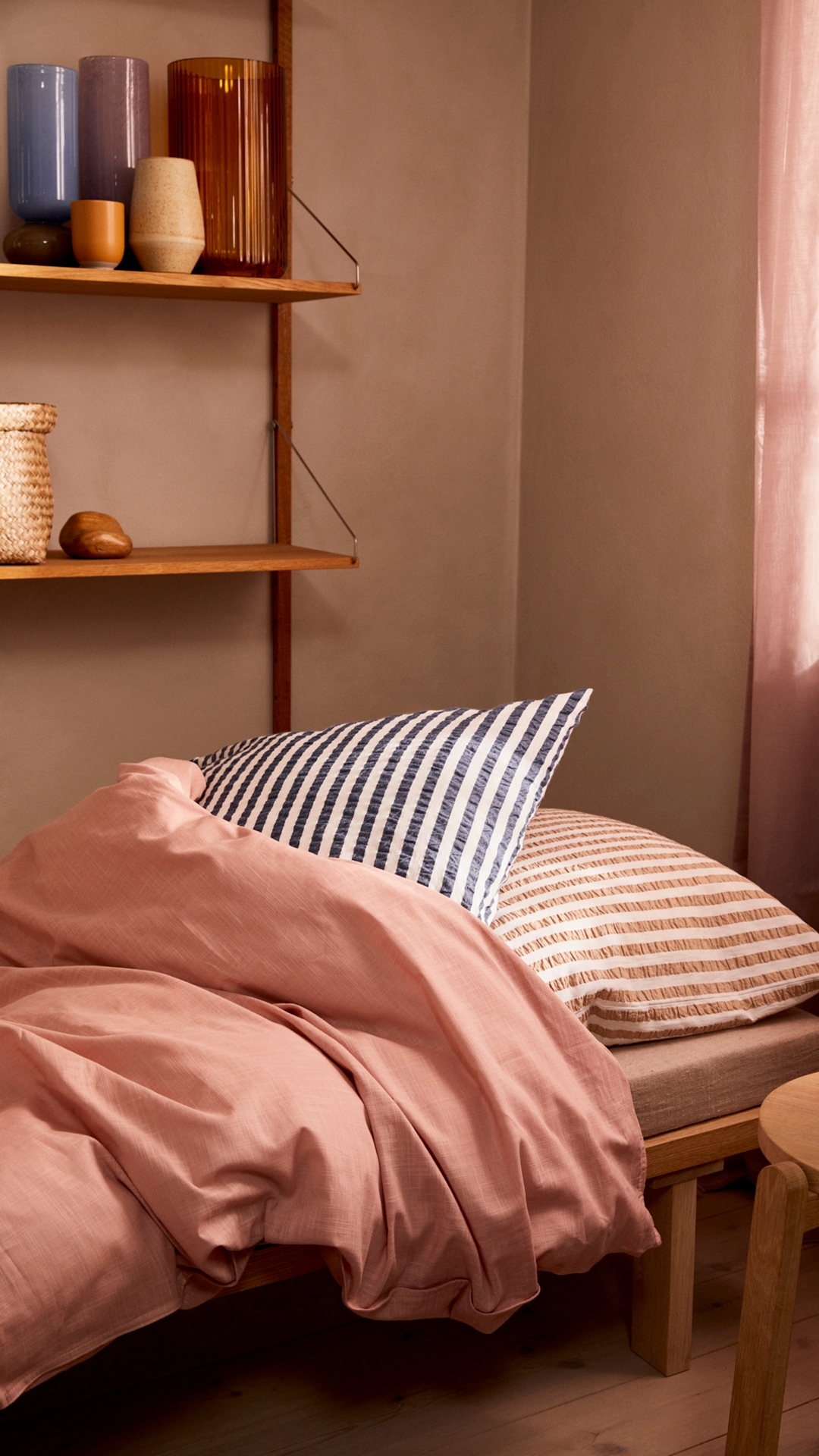 Sovrum med md rosa täcke och randigt rosa kudde, med en trähylla på väggen med vaser på