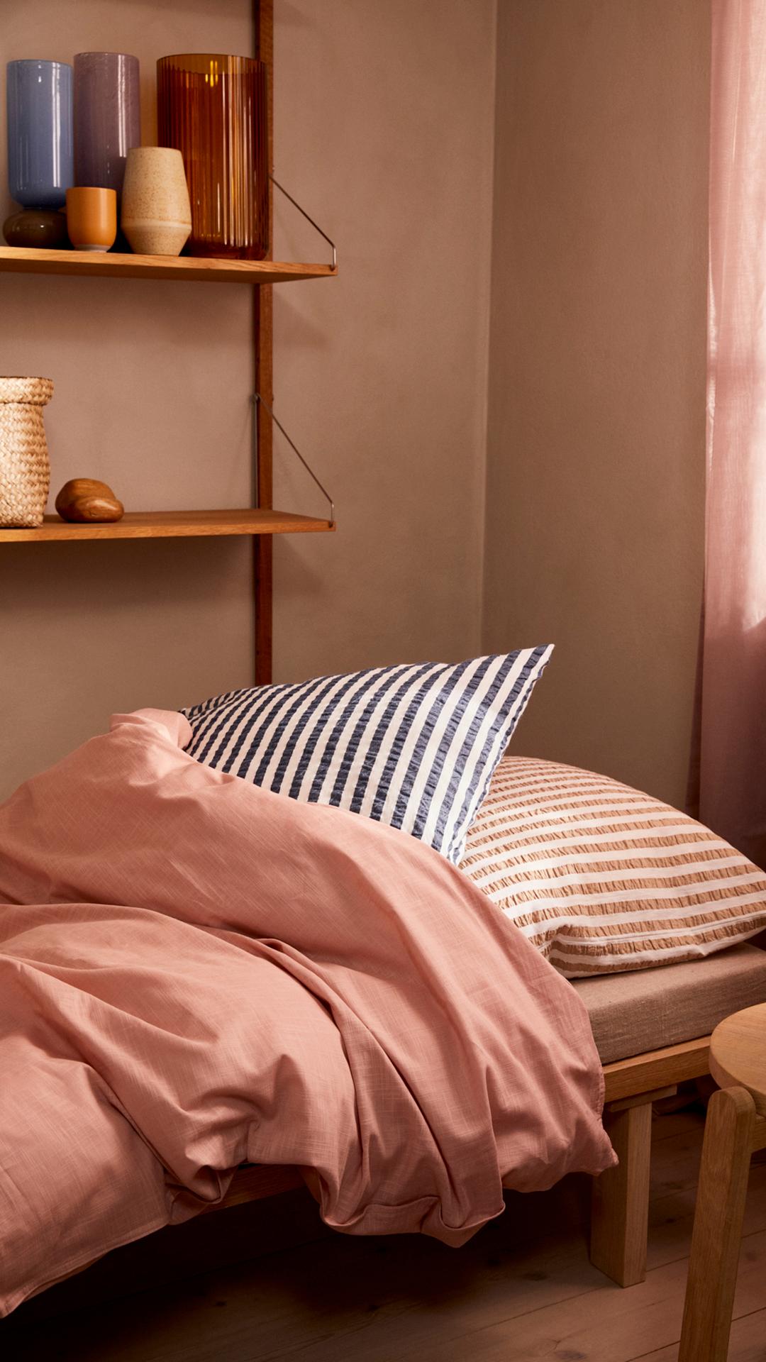 Sovrum med md rosa täcke och randigt rosa kudde, med en trähylla på väggen med vaser på