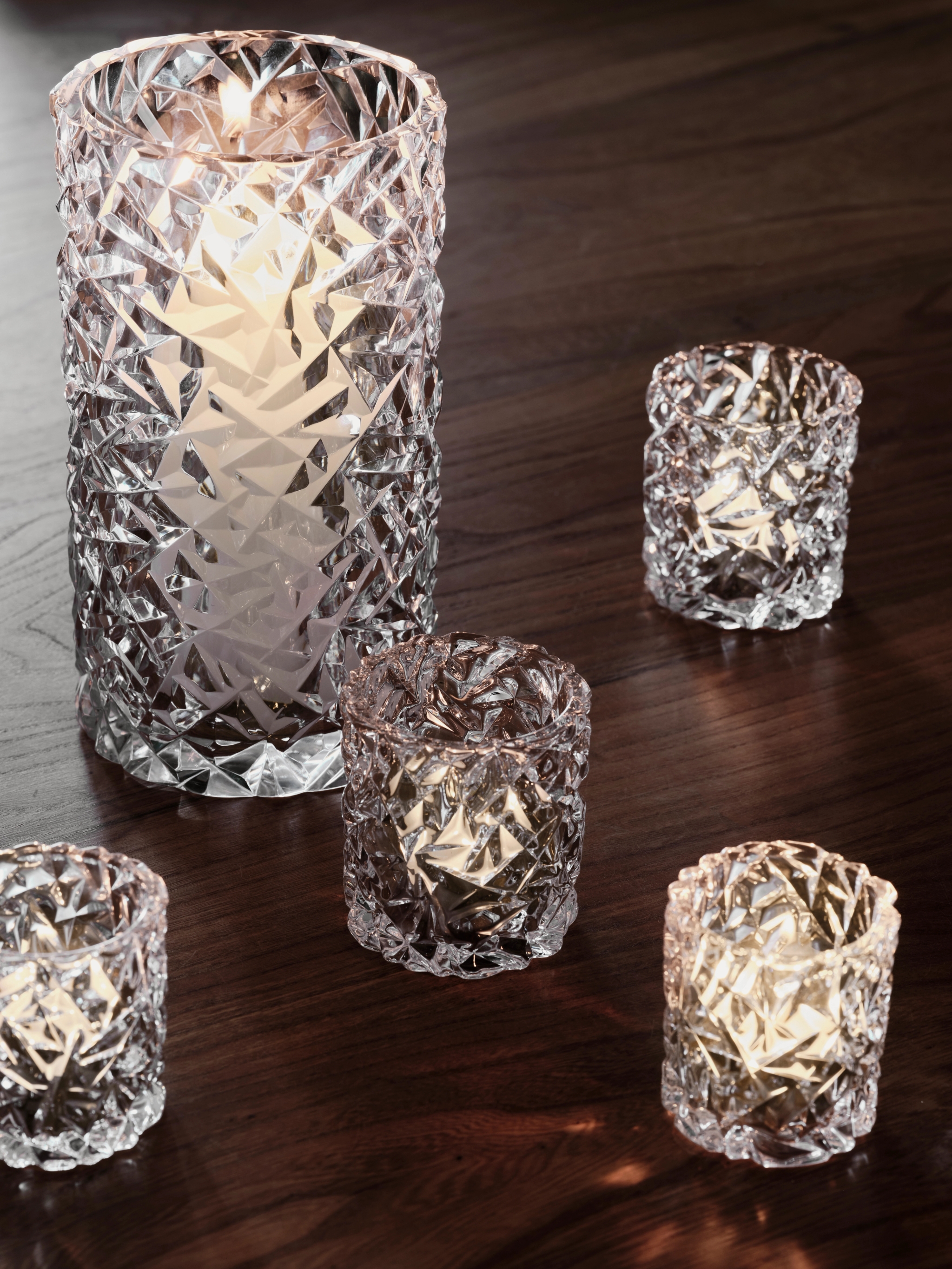 Five clear textured glass candle holders with lit candles on a dark wood surface.