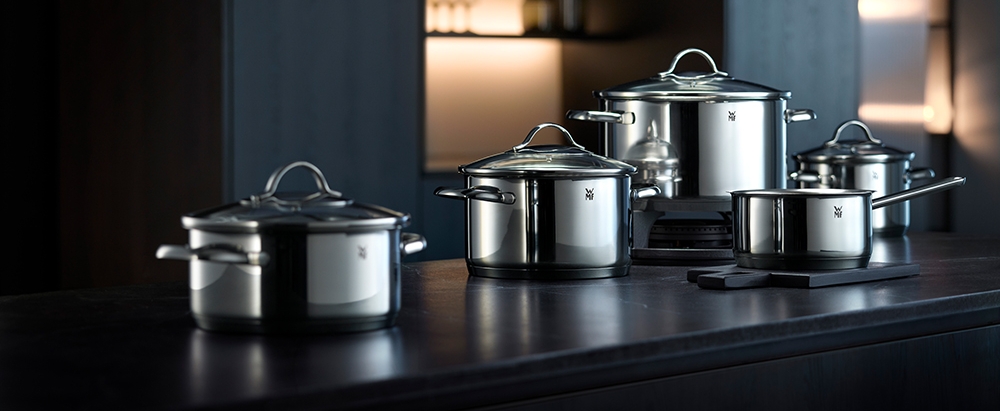 Erilaisia WMF:n ruostumattomasta teräksestä valmistettuja keittokattiloita ja -pannuja tummalla keittiötasolla.