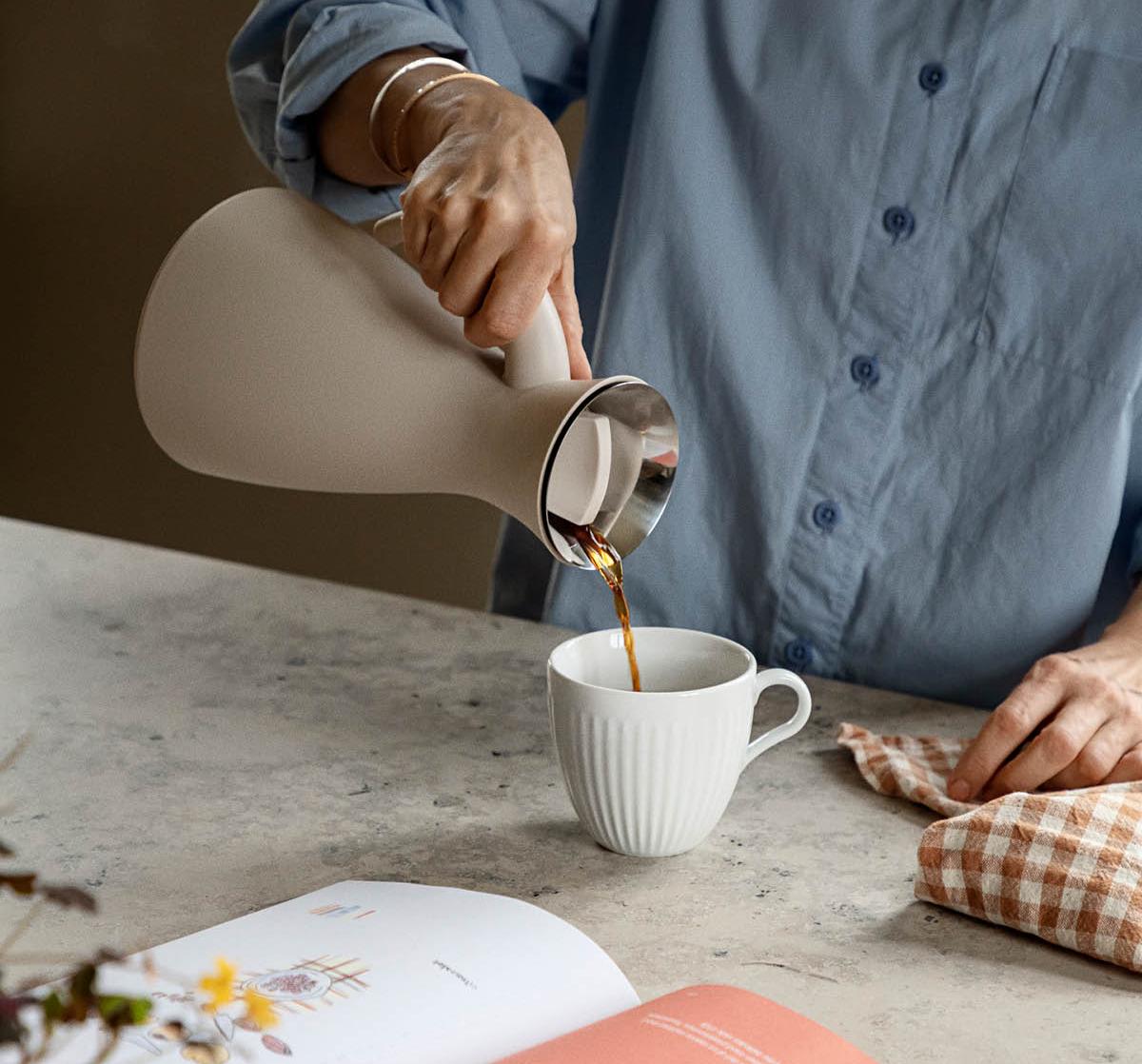 Person häller kaffe ur en stilren beige termoskanna i en vit kopp på ett köksbord.