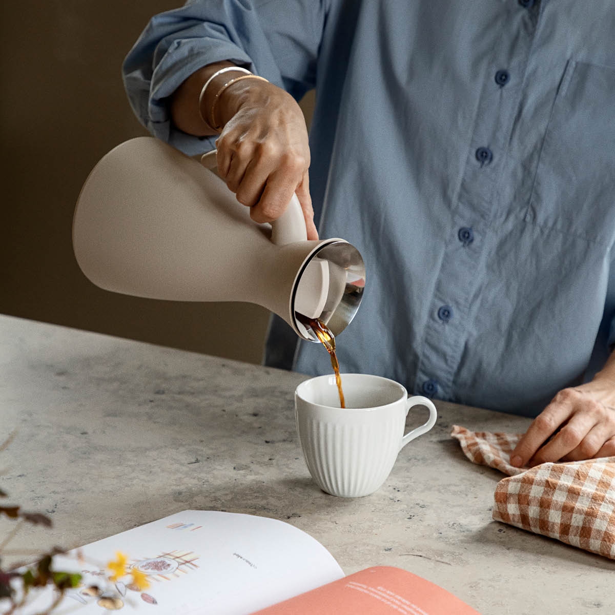Person häller kaffe ur en stilren termoskanna i en vit kopp på ett köksbord.
