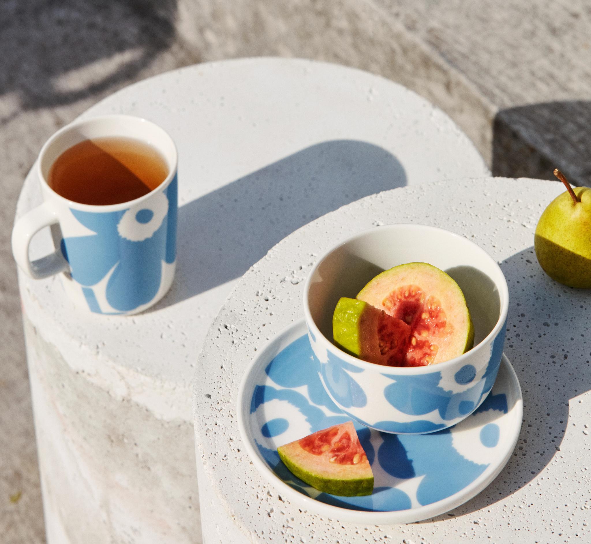 Kesäinen kattaus ulkona: sinikuvioinen muki ja kulho, joissa on teetä ja viipaloitua guavaa, betonipöydällä päärynän vieressä.