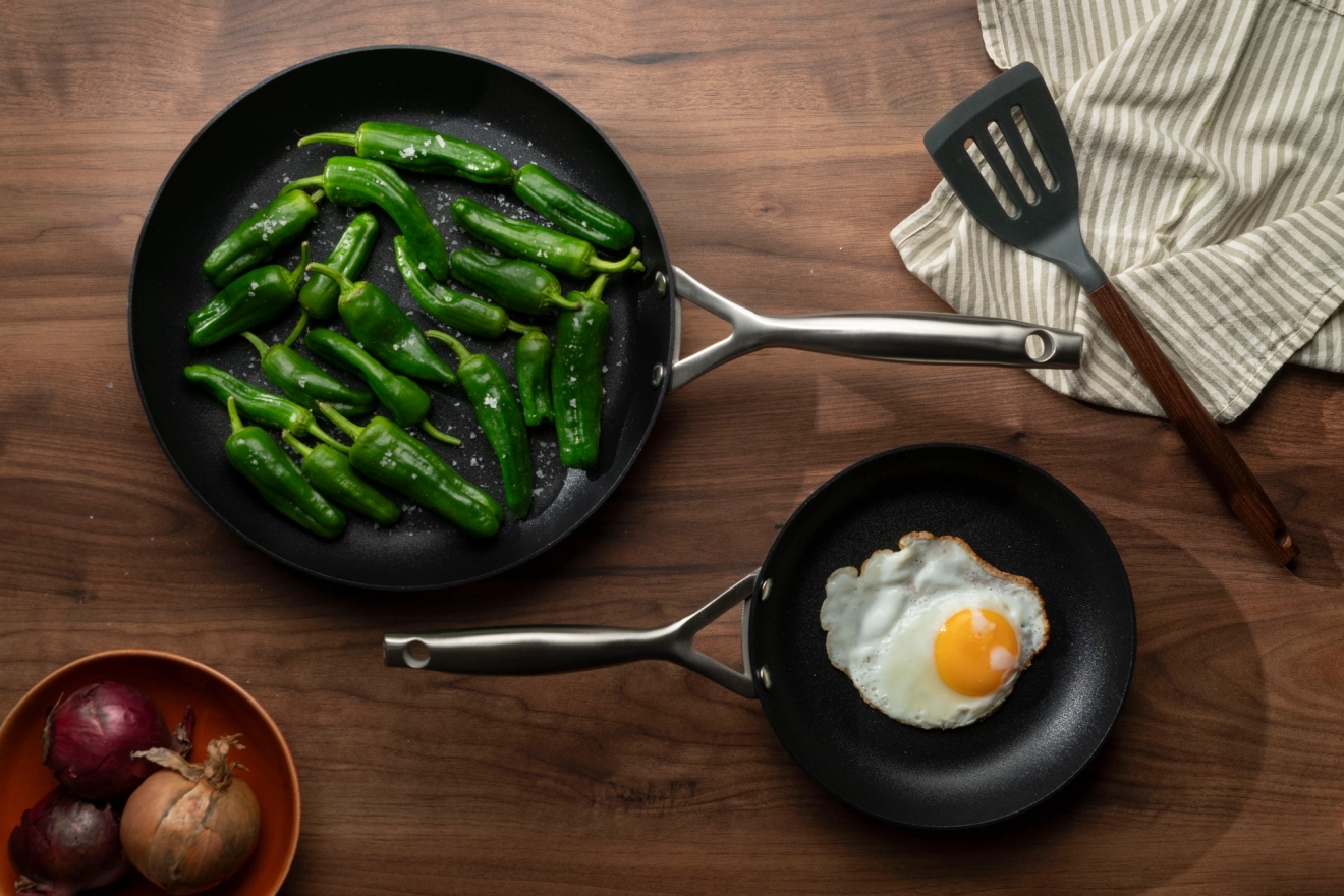 Två stekpannor på ett träbord: en med gröna paprikor, en annan med ett stekt ägg, med en stekspade och lök i närheten.
