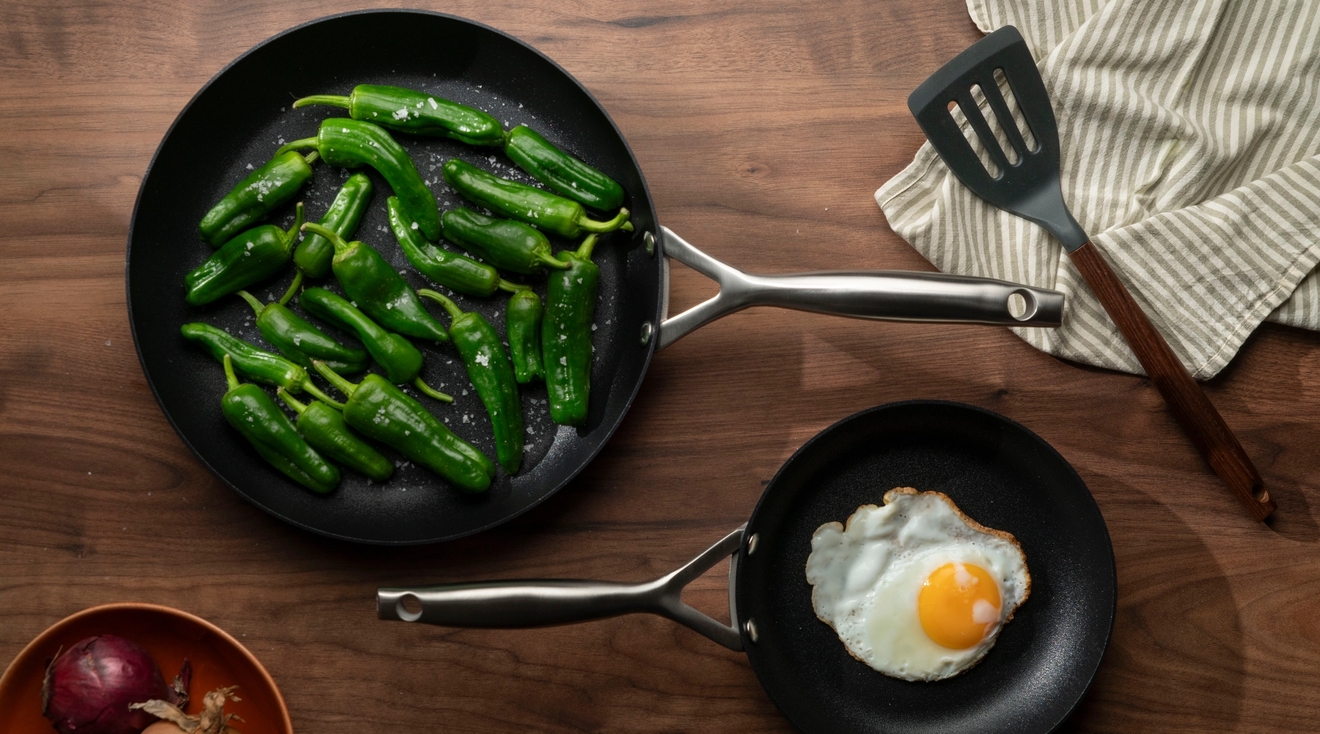Två stekpannor som är fotade uppifrån som står på ett träbord, en med stekt ägg och en med padrons i