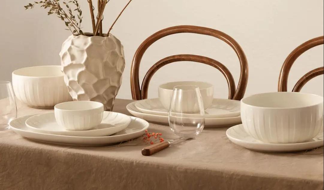 Stilrent dukat matbord med beige linneduk, vita keramikskålar och tallrikar, glas samt en dekorativ vas med torkade blommor framför böjträstolar.