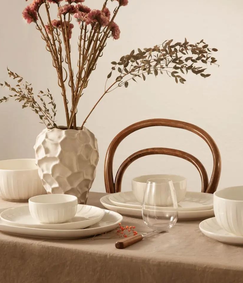 Ett matbord dukat med vitt räfflat porslin, genomskinliga glas och en vit vas med textur med torkade blommor, på en beige duk.