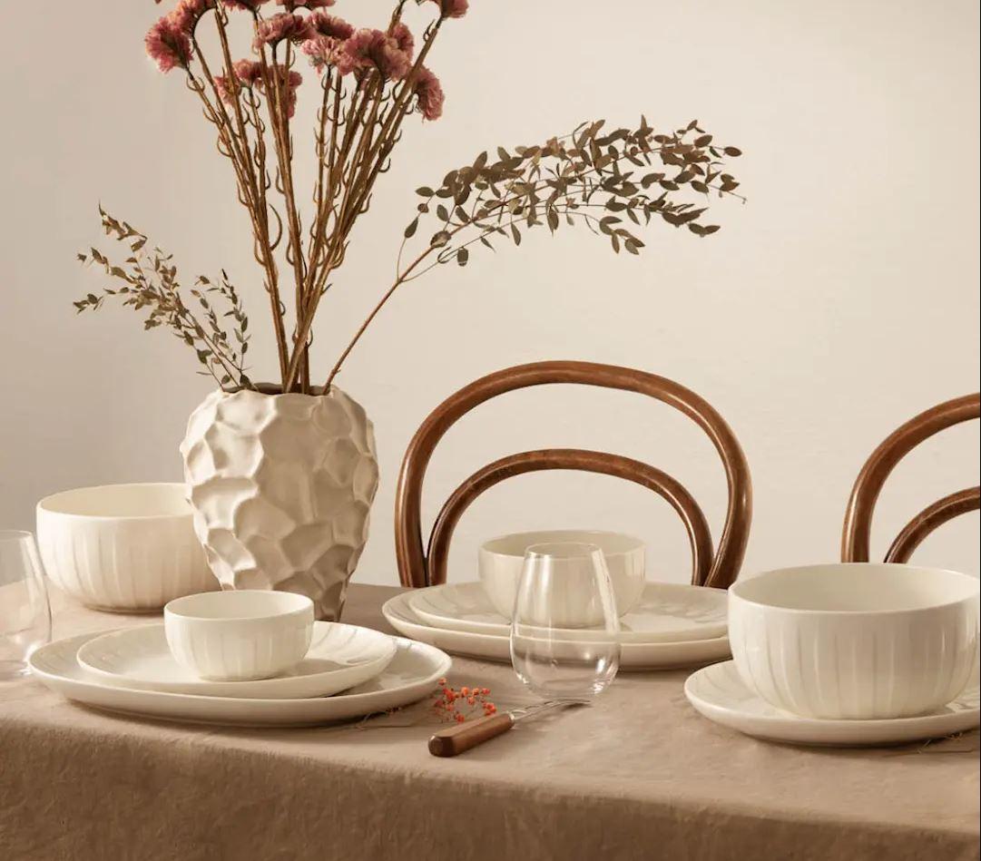 A dining table set with white dishes, a textured vase of dried flowers, and wooden chair backs.