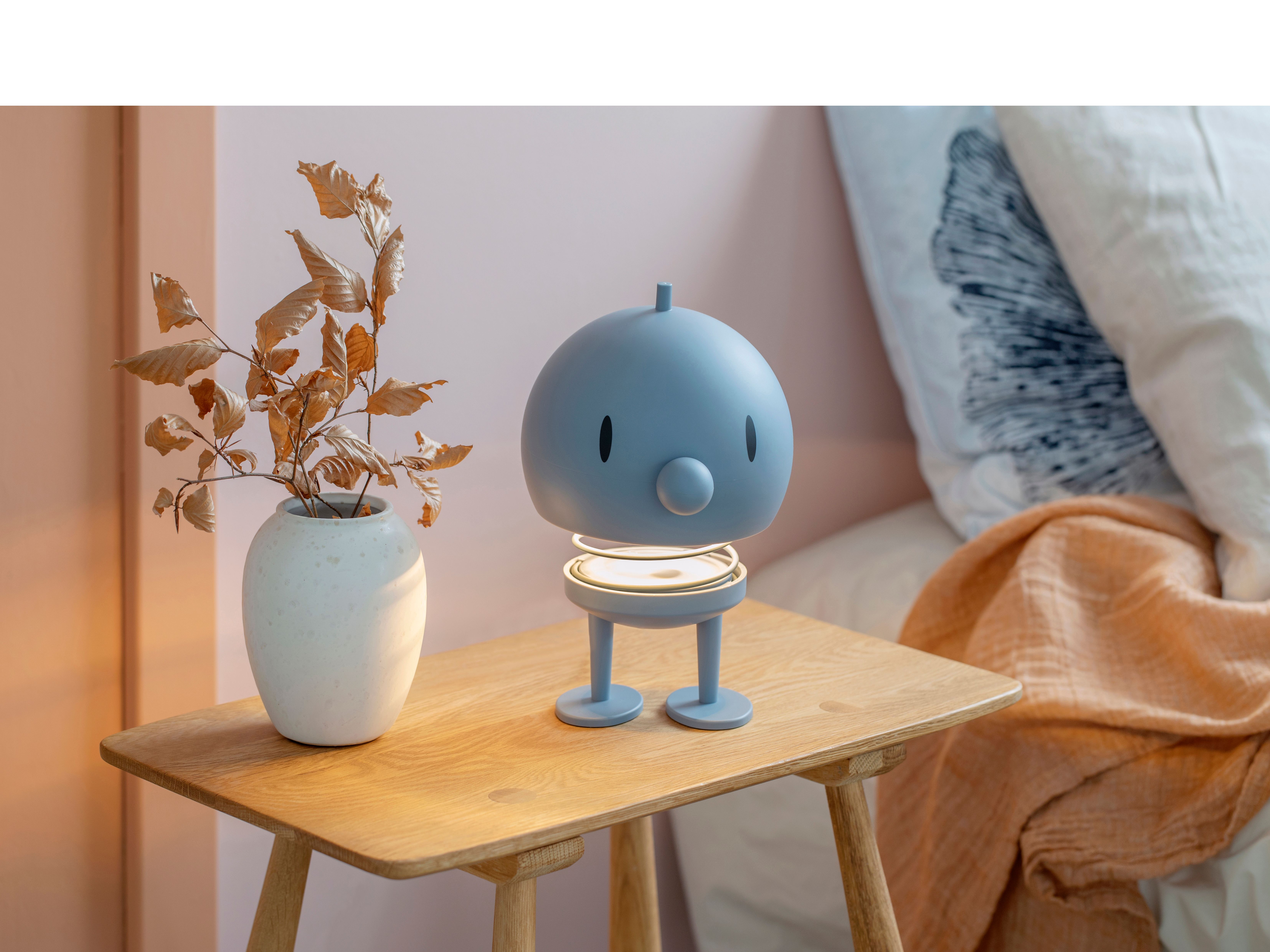 A light blue, character-shaped lamp with a glowing base and a white vase with dried leaves sit on a wooden nightstand.En ljusblå, karaktärsformad lampa med en lysande fot på ett nattduksbord i trä bredvid en vas med torkade blad.