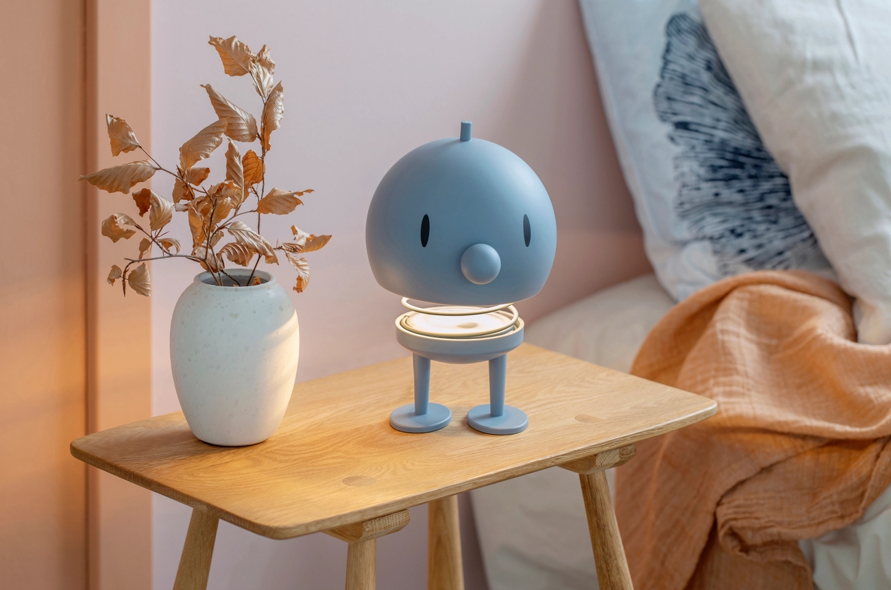 A light blue, character-shaped lamp with a glowing base and a white vase with dried leaves sit on a wooden nightstand.En ljusblå, karaktärsformad lampa med en lysande fot på ett nattduksbord i trä bredvid en vas med torkade blad.