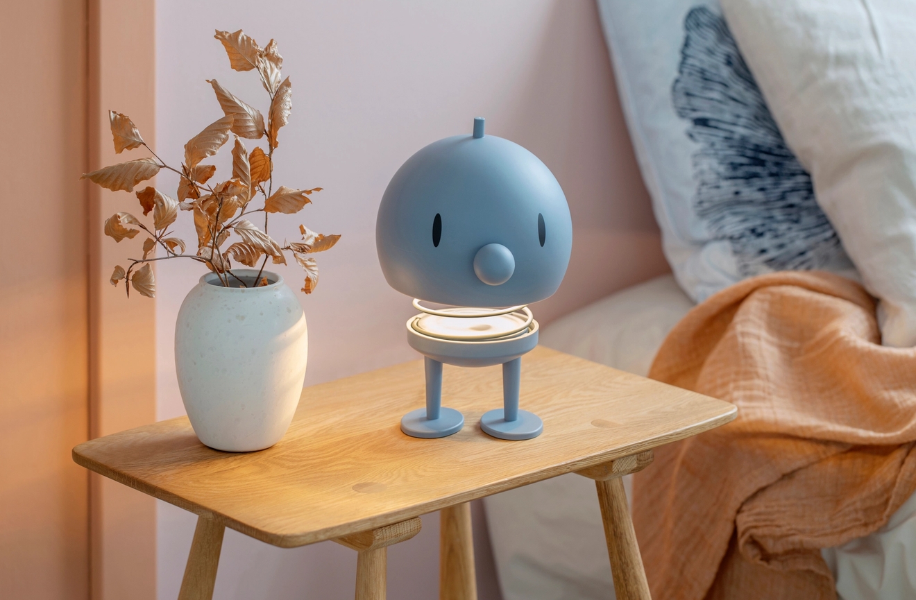 En ljusblå, karaktärsformad lampa med en lysande fot på ett nattduksbord i trä bredvid en vas med torkade blad.