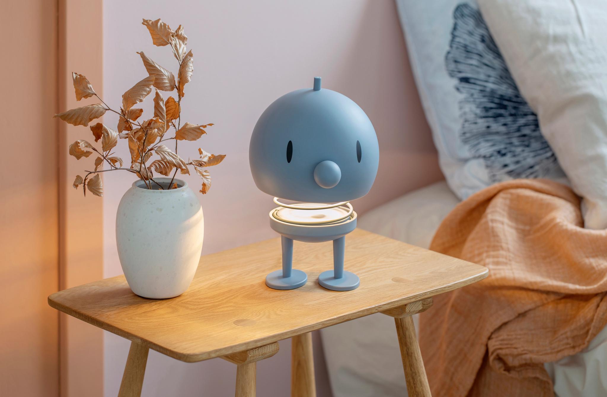 Blå, figurformad bordslampa står på ett träbord bredvid en vit vas med torkade blad i ett mysigt sovrum.