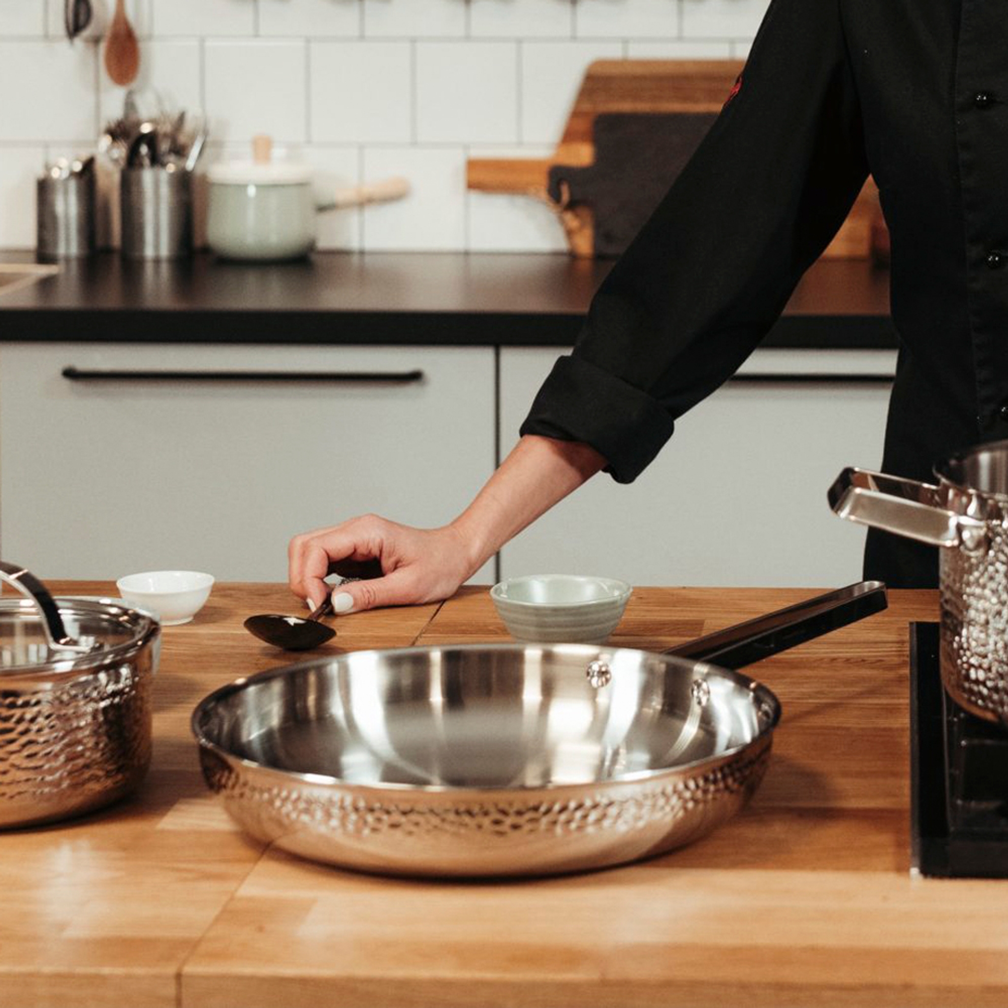 A chef's hand rests on a wooden counter with hammered stainless steel cookware in a kitchen.Vasaroituja kuparikeittovälineitä liedellä keittiössä, jossa on laatoitetut seinät, tuoretta basilikaa ja puisia ruokailuvälineitä.