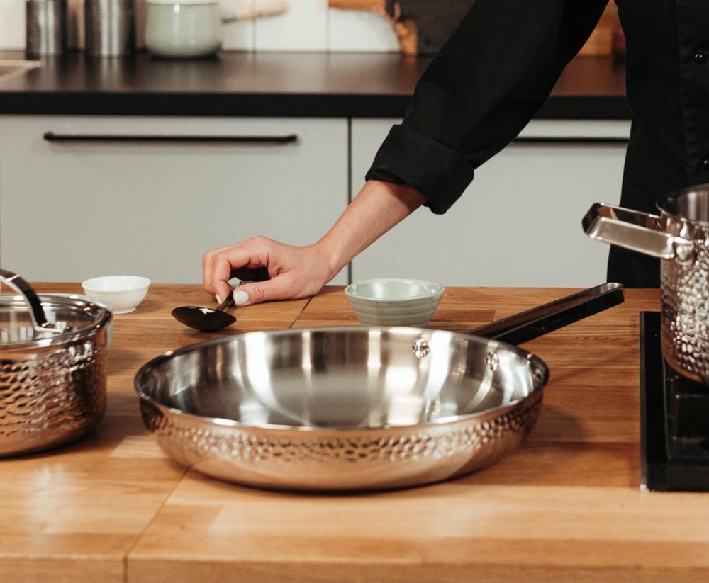 En kockhand håller en sked nära en hamrad stekpanna i rostfritt stål på en träbänk i ett kök.