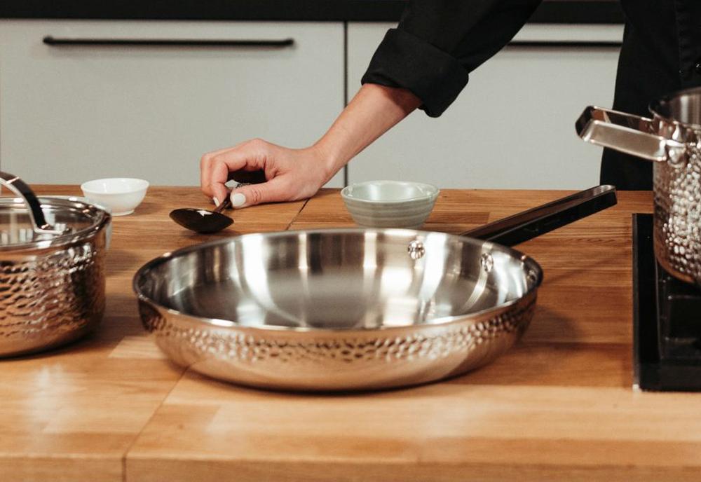 En kockhand håller en sked nära en hamrad stekpanna i rostfritt stål på en träbänk i ett kök.