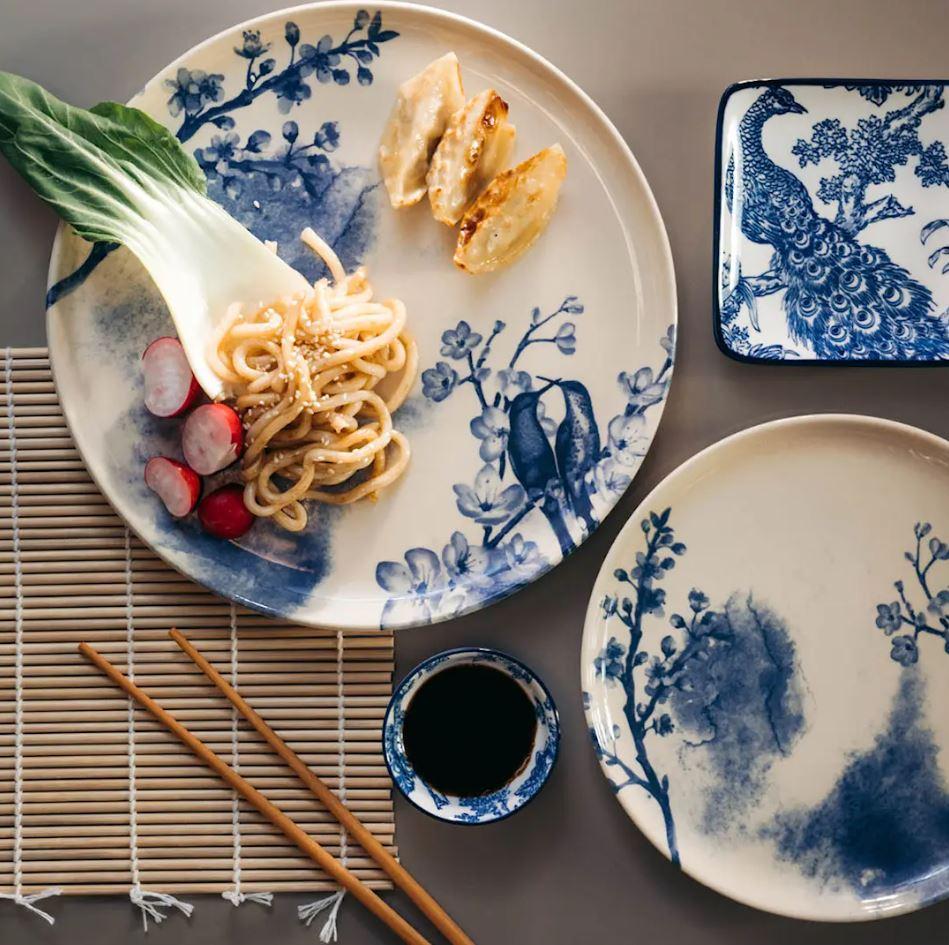 Overhead shot of an Asian meal with noodles, bok choy, radishes, and dumplings on white plates with blue oriental bird and floral designs, alongside soy sauce, chopsticks, and a bamboo mat.