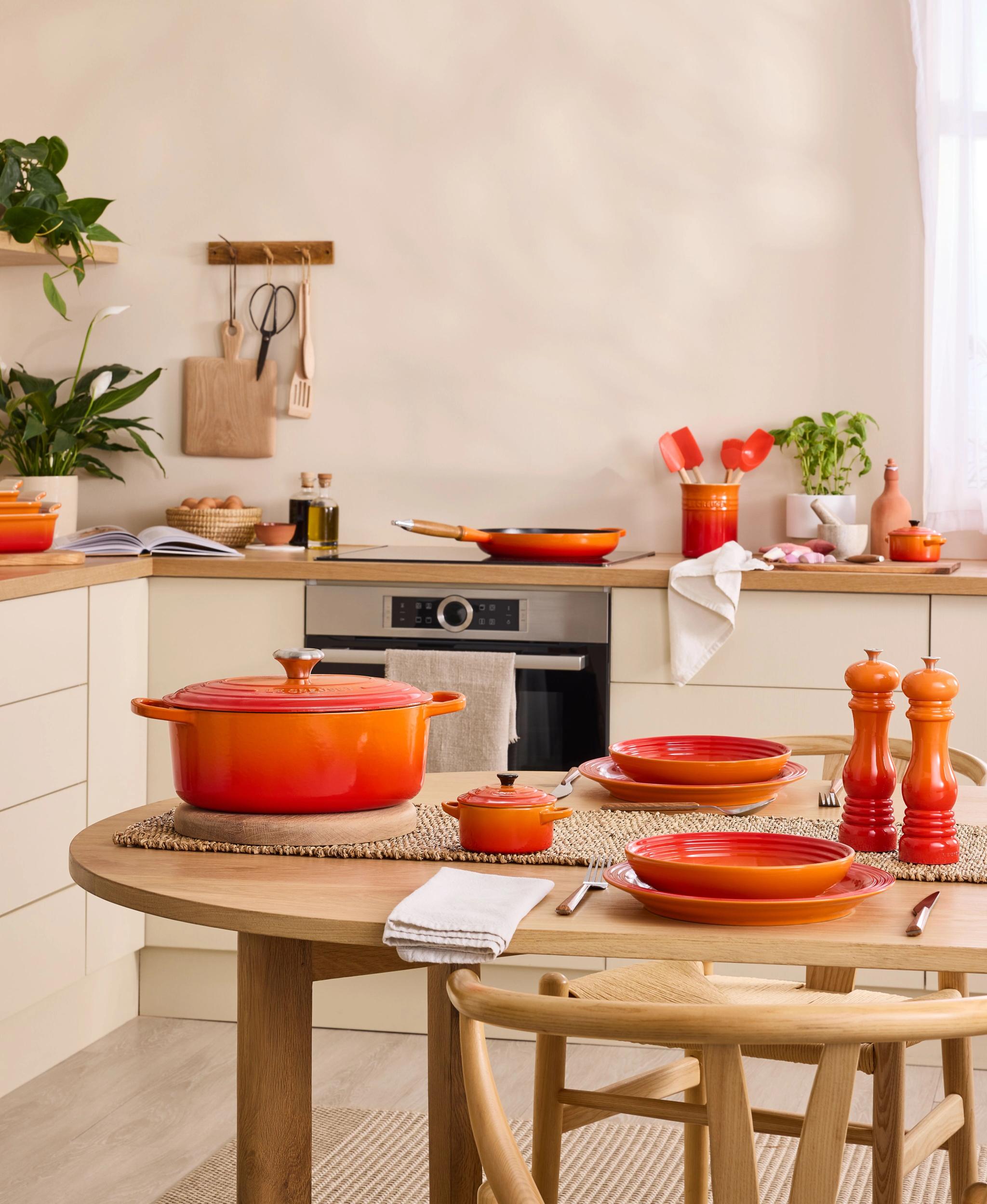 Et lyst kjøkken med et trebord som det står en oransje støpejernsgryte og oransje tallerkener på