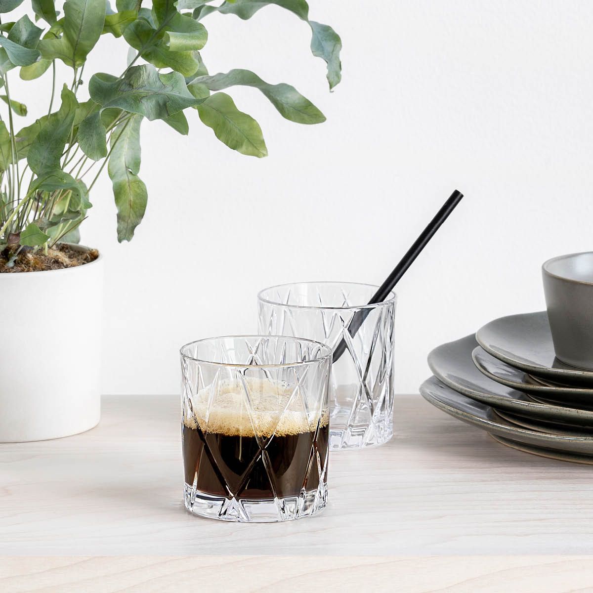 Stilren dukning med två kristallglas med kaffe, en grön krukväxt och staplade tallrikar på ett ljust träbord.