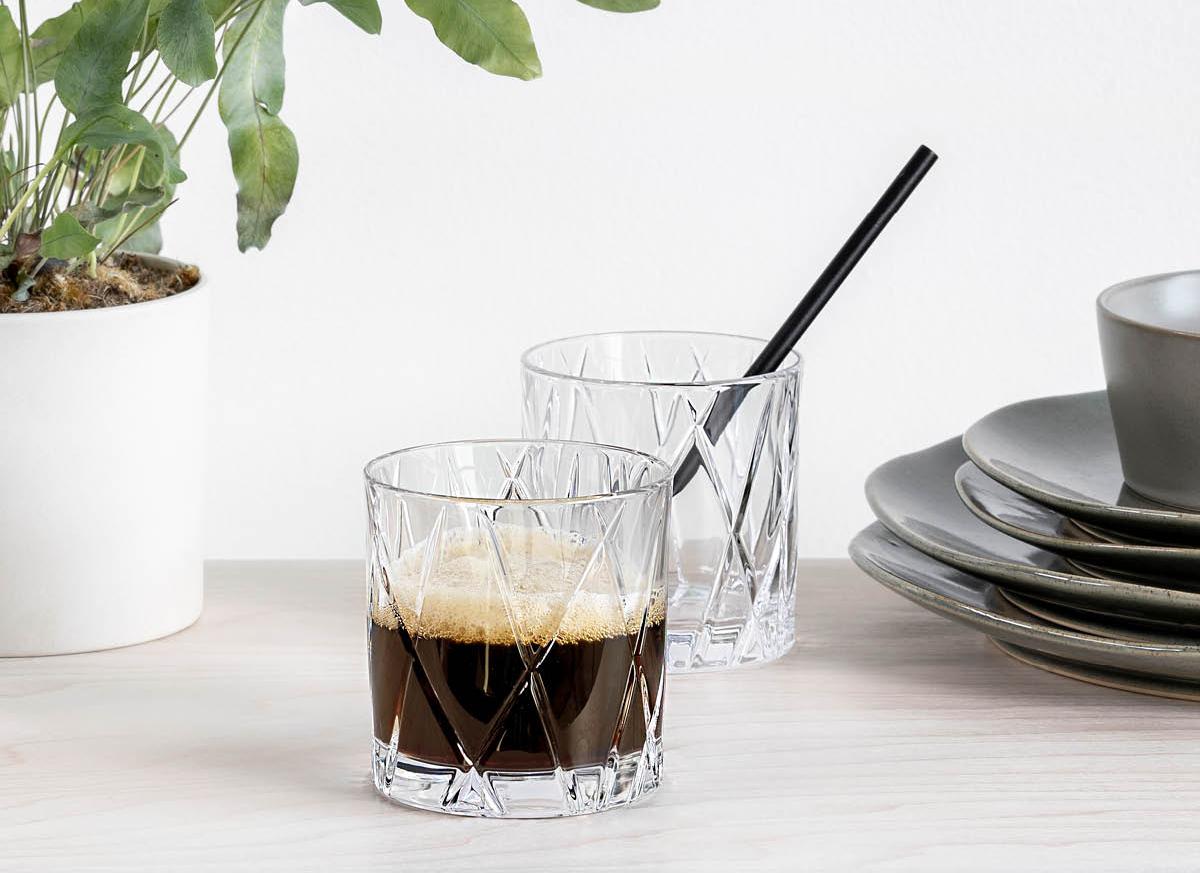 Två kristallglas med kaffe står på ett ljust bord bredvid en grön krukväxt och en stapel tallrikar.