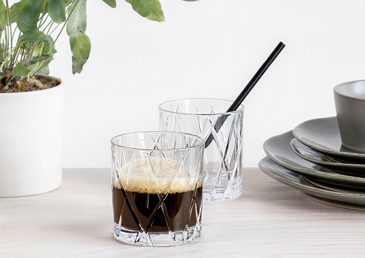 Två eleganta glas med mörk dryck står på ett ljust bord bredvid en grön krukväxt och staplade tallrikar.
