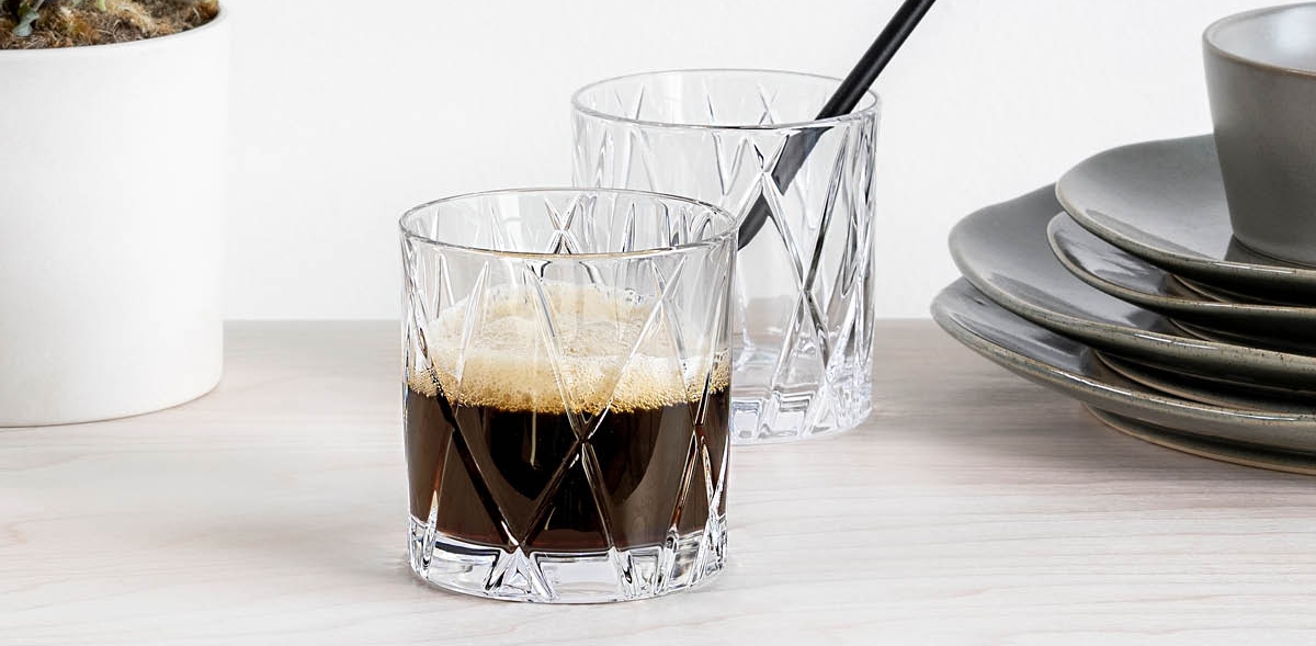 Stilren dukning med två kristallglas med kaffe, en grön krukväxt och staplade tallrikar på ett ljust träbord.