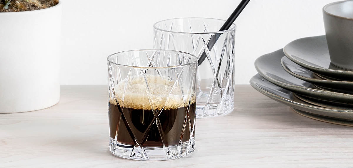 Två eleganta kristallglas med kaffe står på ett ljust bord bredvid en grön krukväxt och staplade tallrikar.