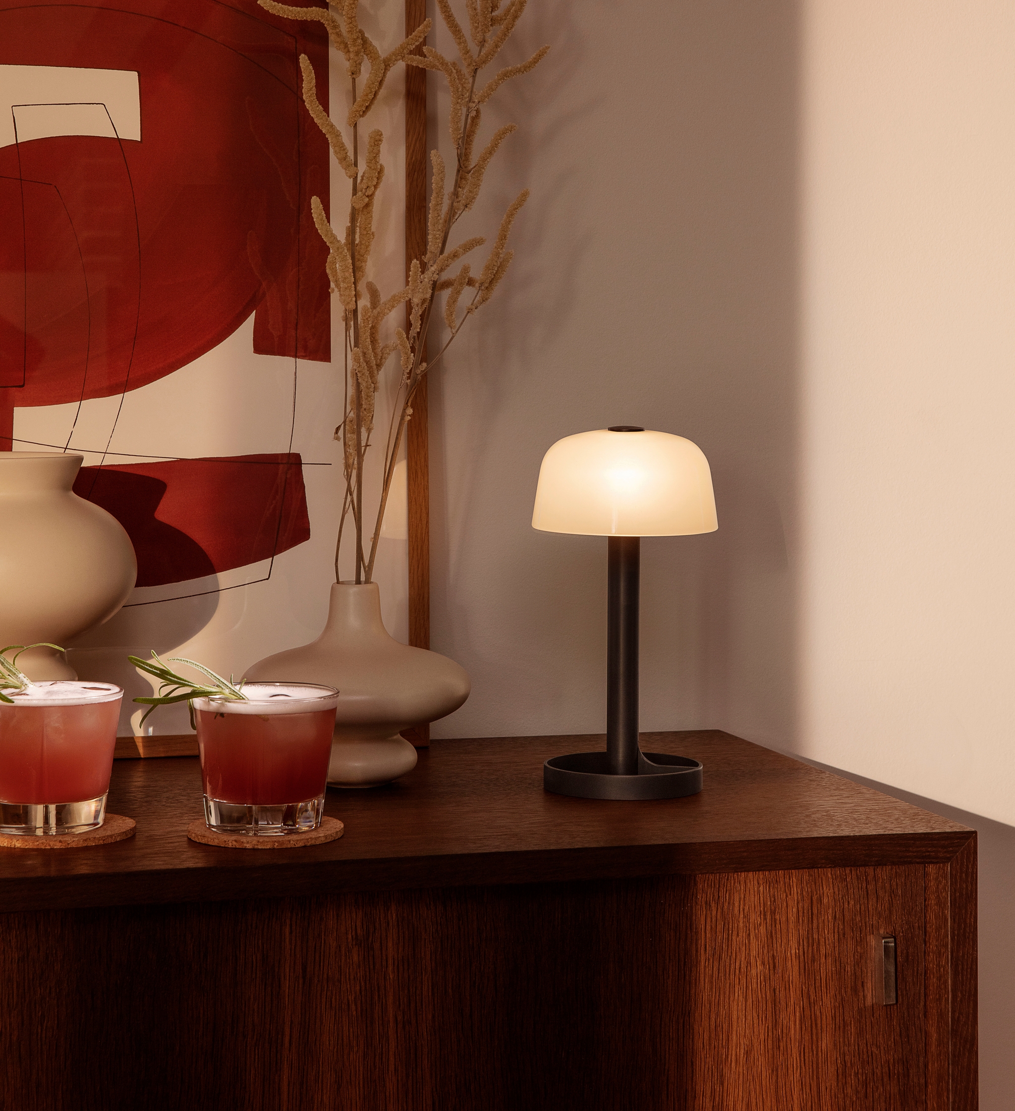 Två cocktails, dekorativa vaser, abstrakt konst och en lysande bordslampa på ett träskåp.