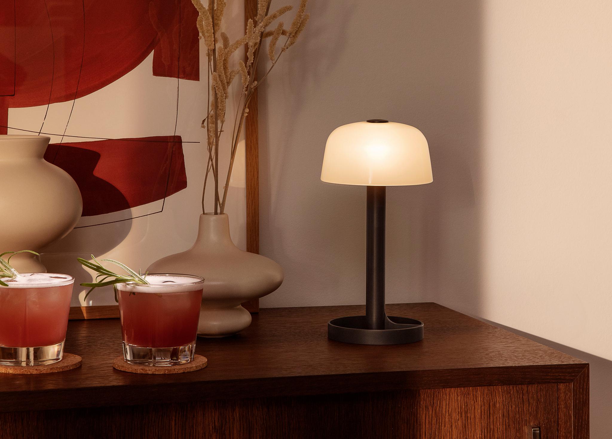 En modern bärbar lampa med en glödande frostad kupol står på ett träskåp, bredvid två röda cocktails, dekorativa vaser och abstrakt konst.