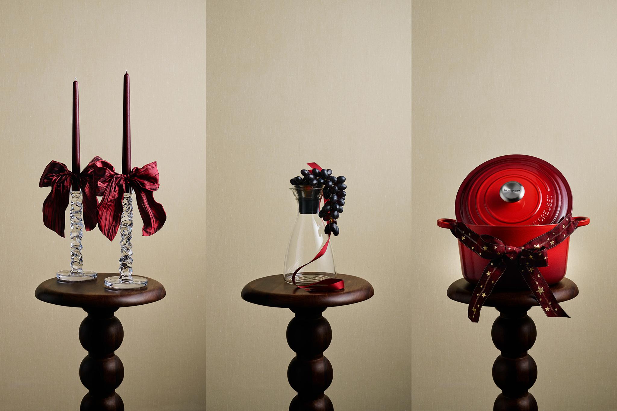Kolme juhlavaa lahjaesinettä jalustoilla: punaiset kynttilät ruseteilla, punainen Le Creuset -ruukku rusetilla ja pinotut hopeiset ruukut punaisella nenäliinalla.