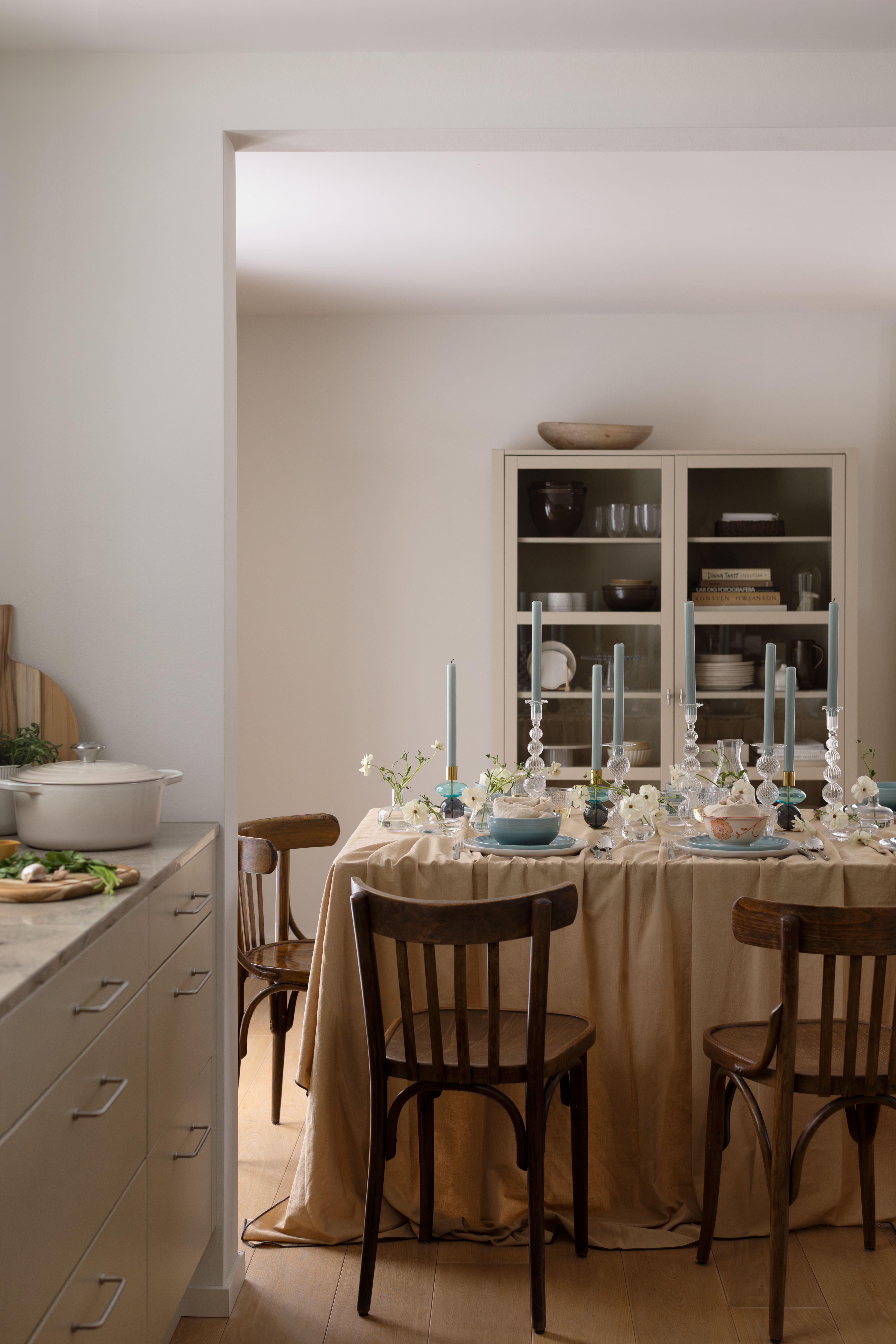 Ett matbord med en beige duk, blå ljus och vita blommor, bredvid en köksbänk.