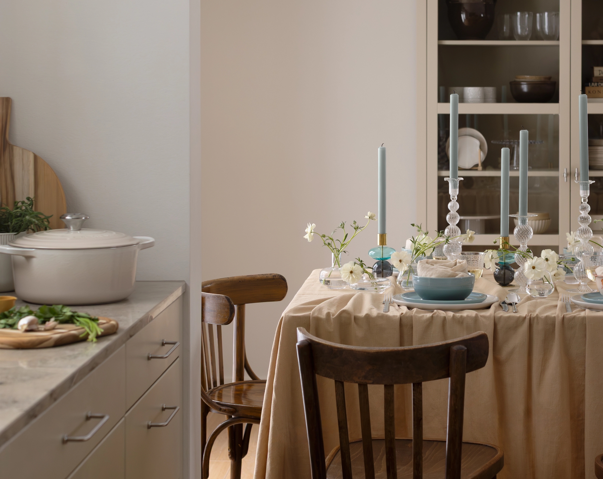 Ett matbord dukat med en beige duk, blå tallrikar, vita blommor och höga blå ljus, med ett kök synligt i bakgrunden.