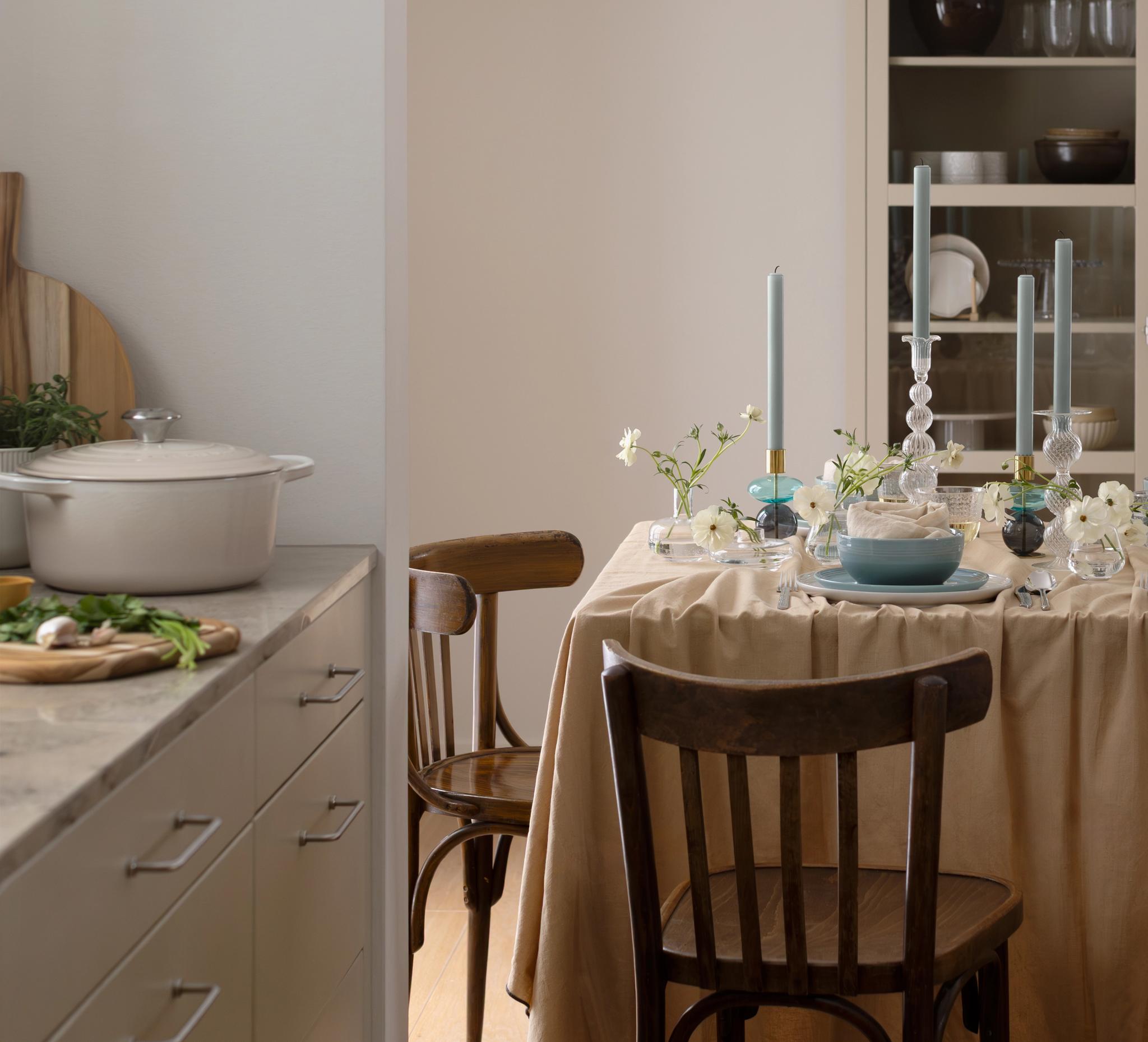 Ett matbord dukat med en beige duk, blå tallrikar, vita blommor och höga blå ljus, med ett kök synligt i bakgrunden.