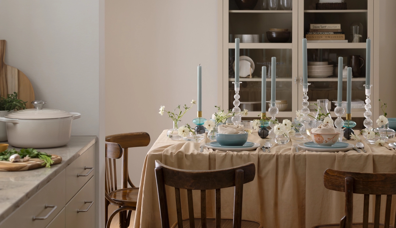 Ett matbord dukat med blå ljus, vita blommor och servis, bredvid en köksbänk med en kastrull.