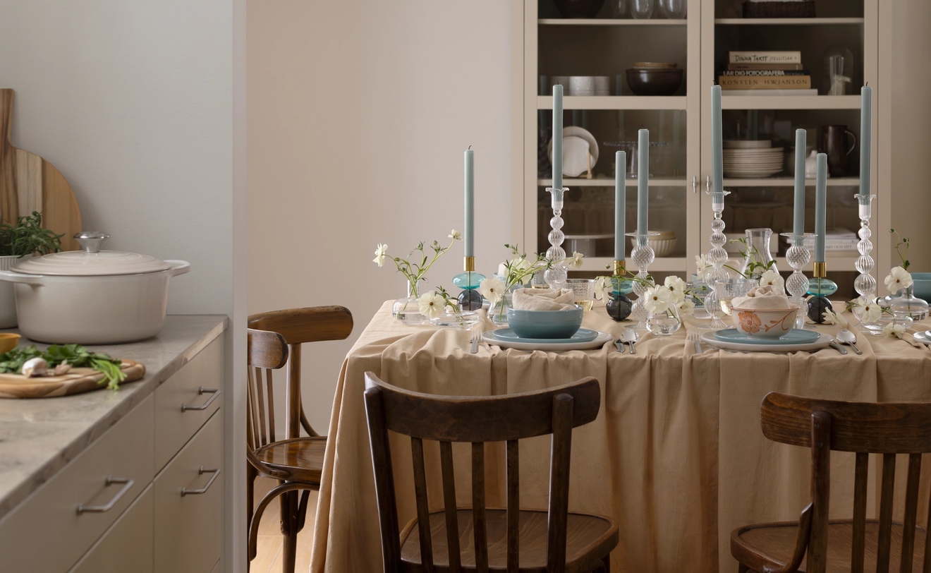 Ett matbord dukat med en beige duk, blå tallrikar, vita blommor och höga blå ljus, med ett kök synligt i bakgrunden.